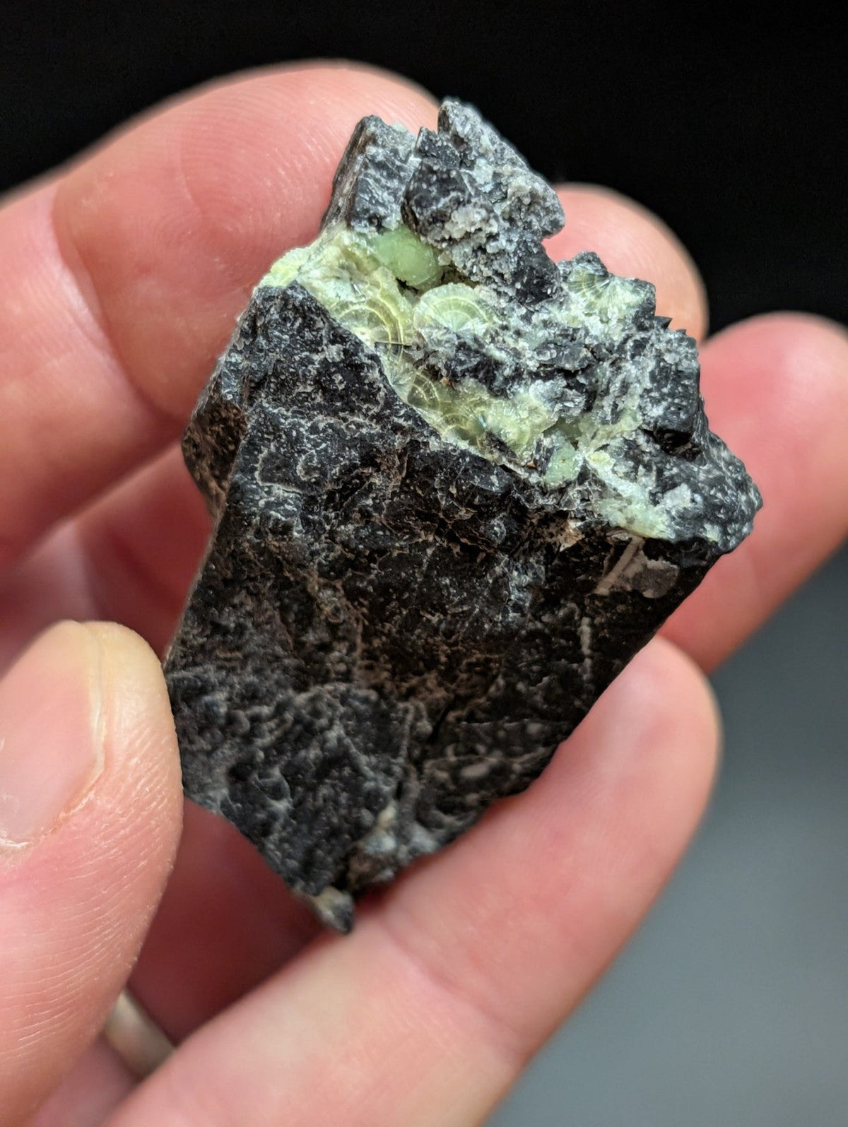 A dark, rough-textured rock with a patch of pale green mineral formations on its surface.