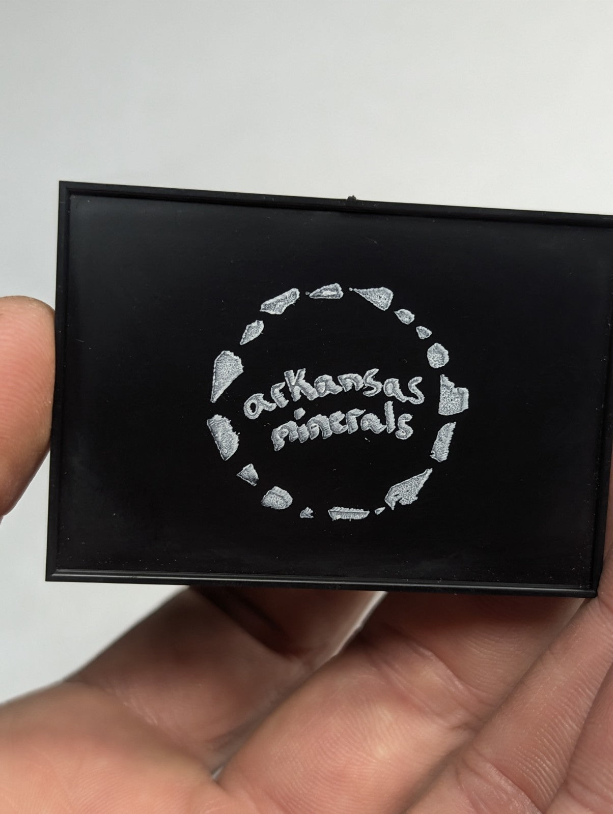 Black rectangular compact with silver embossed ’arkansas minerals’ logo encircled by a ring of stylized mineral fragments.