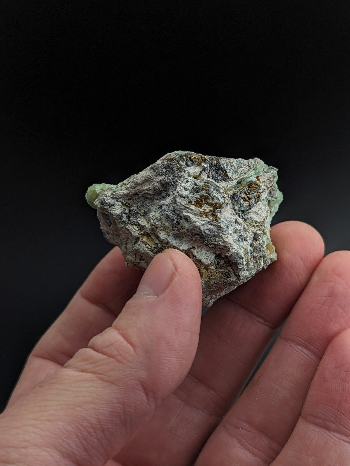 A rough, irregularly shaped mineral specimen with a mottled green and gray surface, featuring patches of earthy brown.