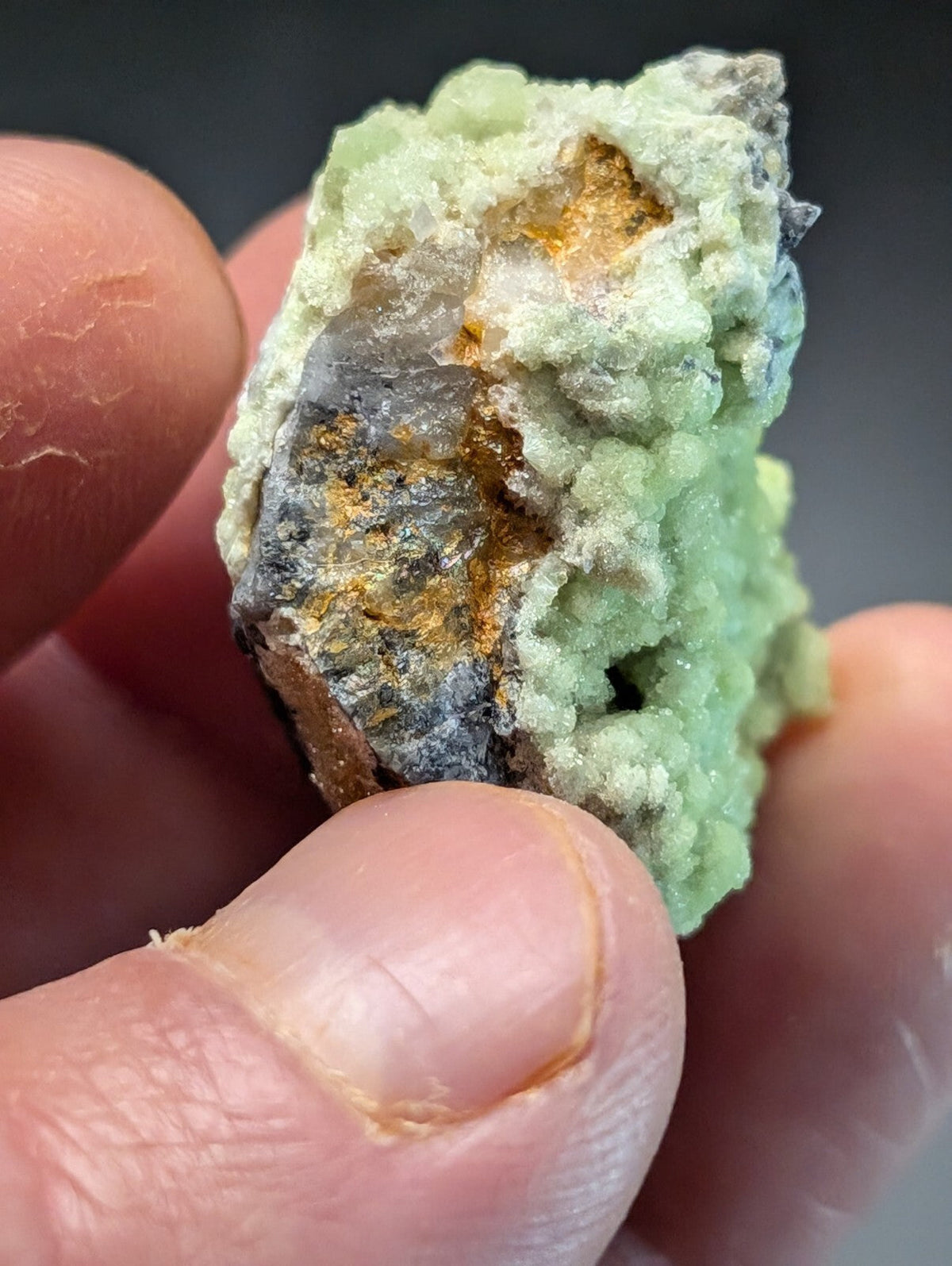 A rough, irregularly shaped mineral specimen with a pale green crystalline exterior and patches of dark gray rock interspersed with golden-yellow inclusions.