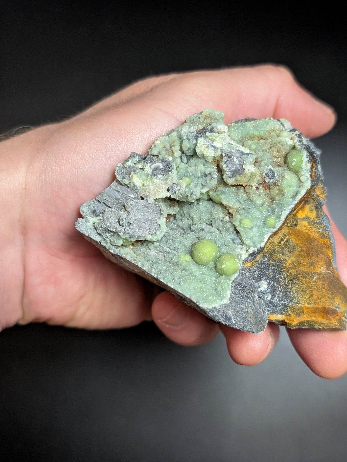 A hand holds a geological specimen featuring vibrant green spherical crystals embedded in a textured matrix of gray and orange mineral formations.
