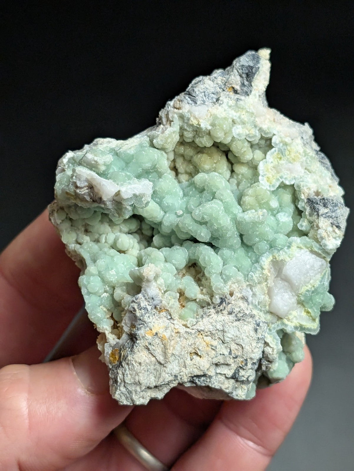 A hand holds a rough, irregularly shaped mineral specimen featuring clusters of pale green, bead-like crystals growing from a grayish matrix.