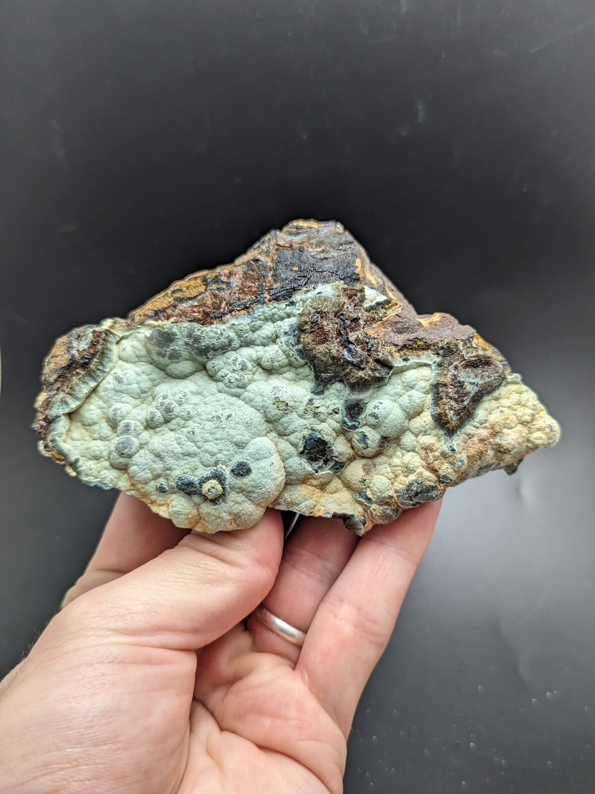 A hand holds a textured, irregularly shaped mineral specimen with patches of pale greenish-blue and brownish-orange hues.
