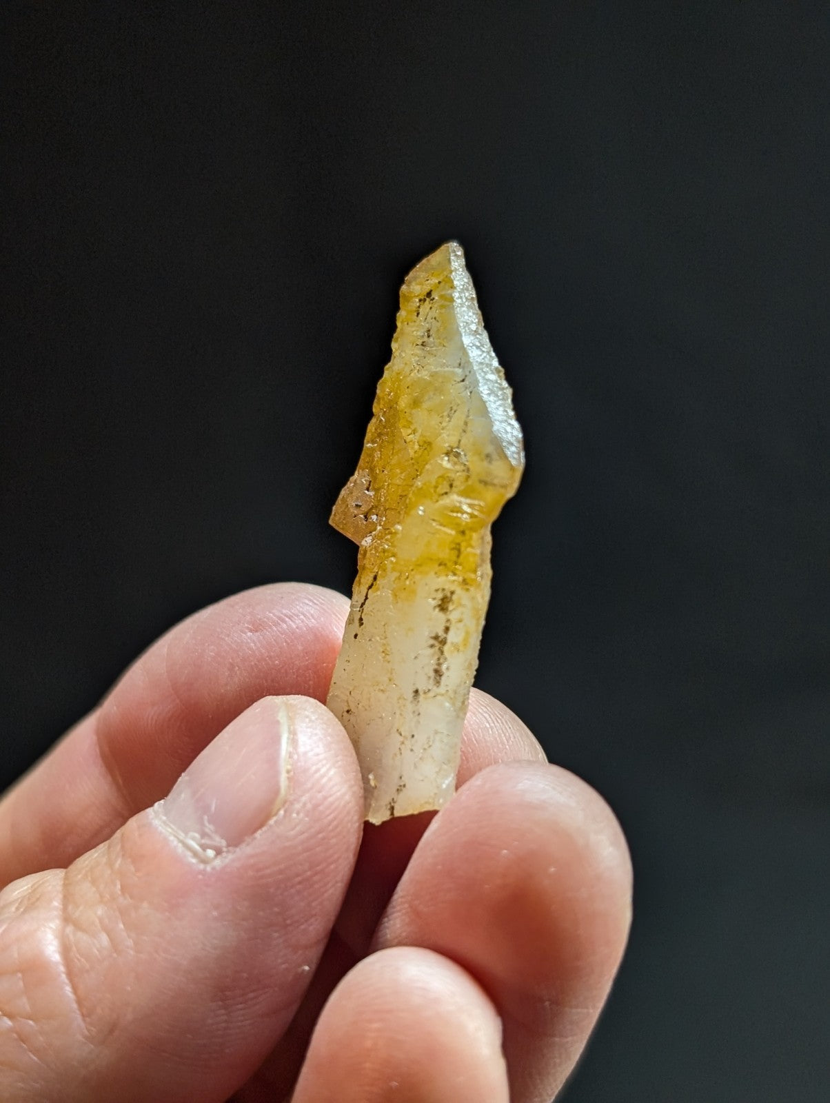 A translucent yellowish crystal with a pointed tip and natural striations, held gently between fingers.