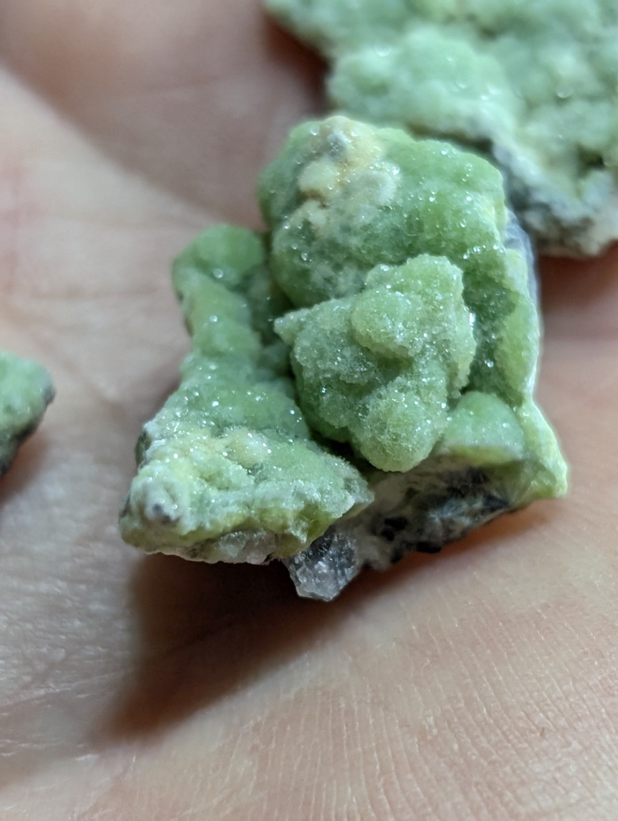 A cluster of light green, crystalline minerals with a rough, textured surface and some darker grayish patches.