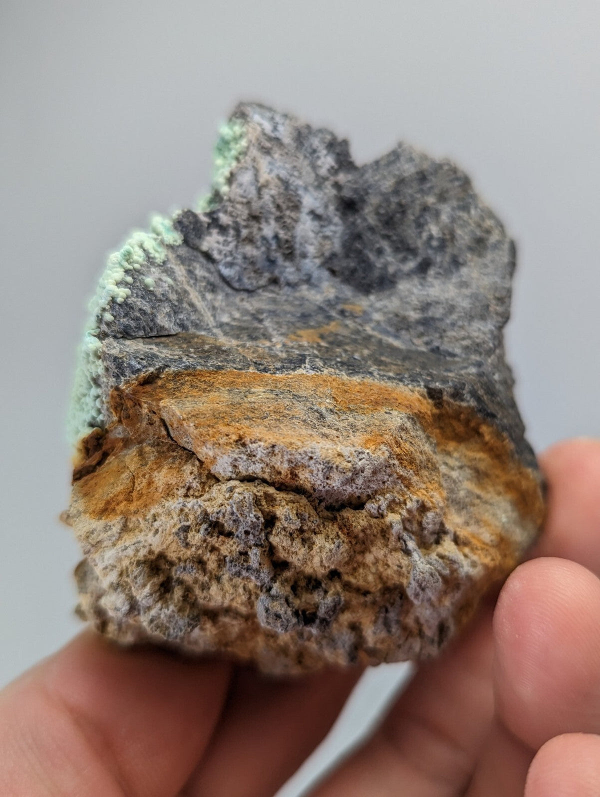 A rugged, multi-toned mineral specimen with a grayish base, orange-brown streaks, and patches of green crystalline growth.