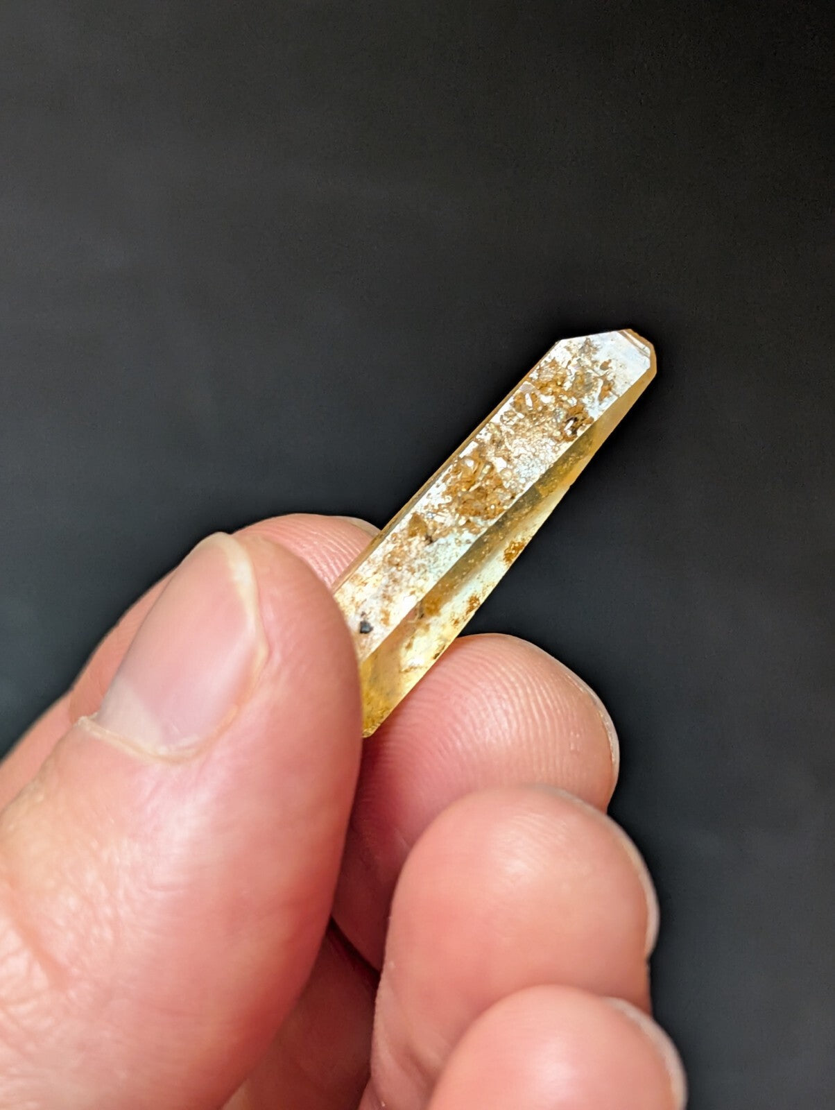 A translucent yellow crystal with golden inclusions and a pointed tip, held gently between fingers.