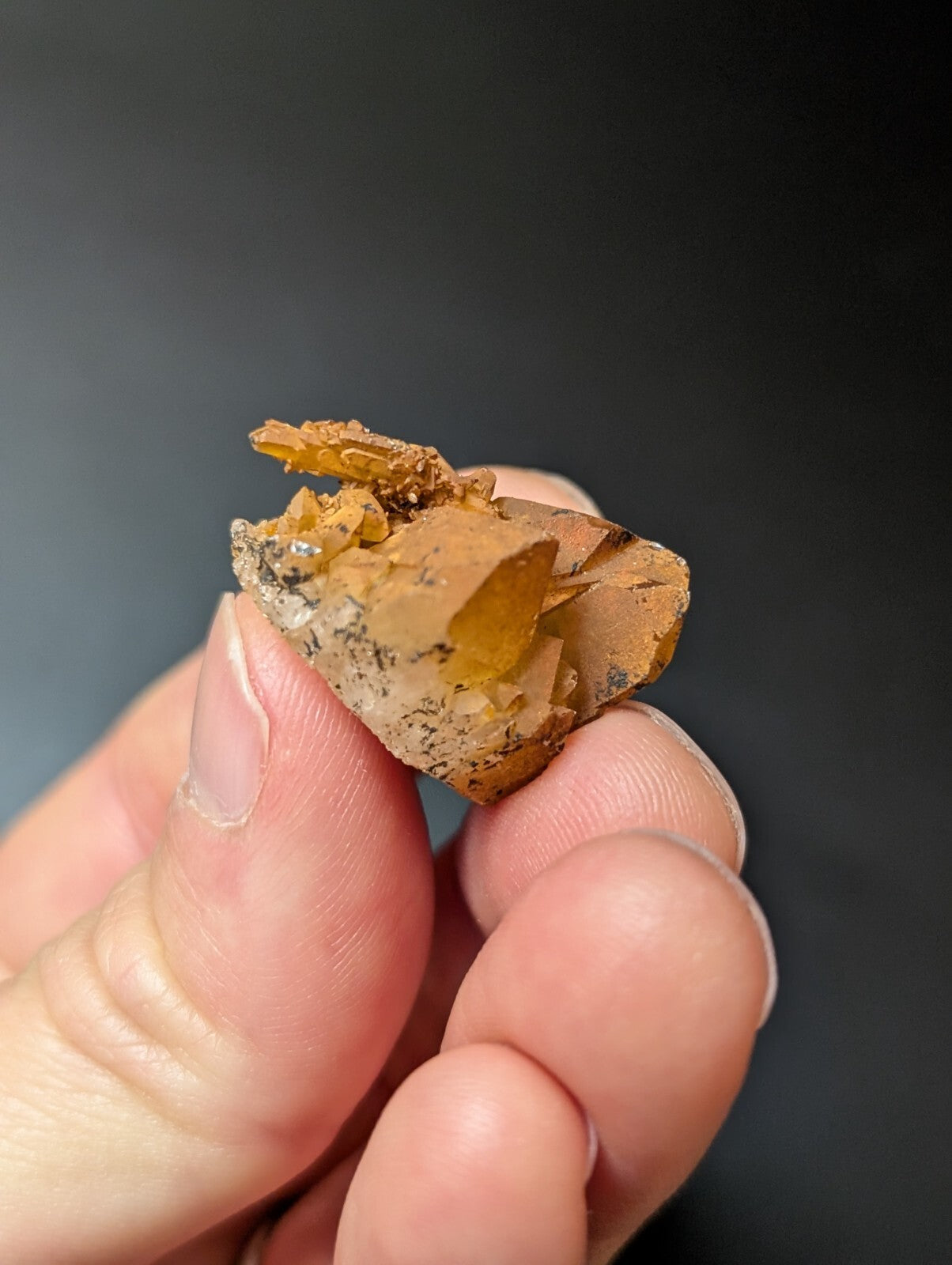 A hand holds a small, irregularly shaped mineral specimen featuring translucent amber-yellow crystals growing from a darker brownish matrix.