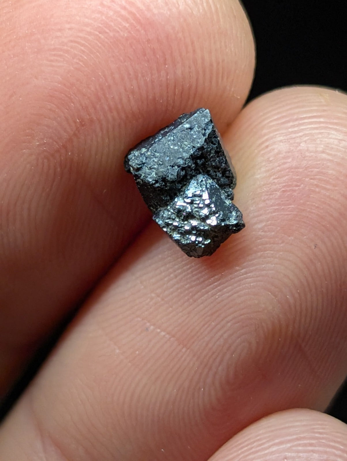 A small, dark metallic mineral specimen with a rough, faceted surface and subtle iridescent sheen.