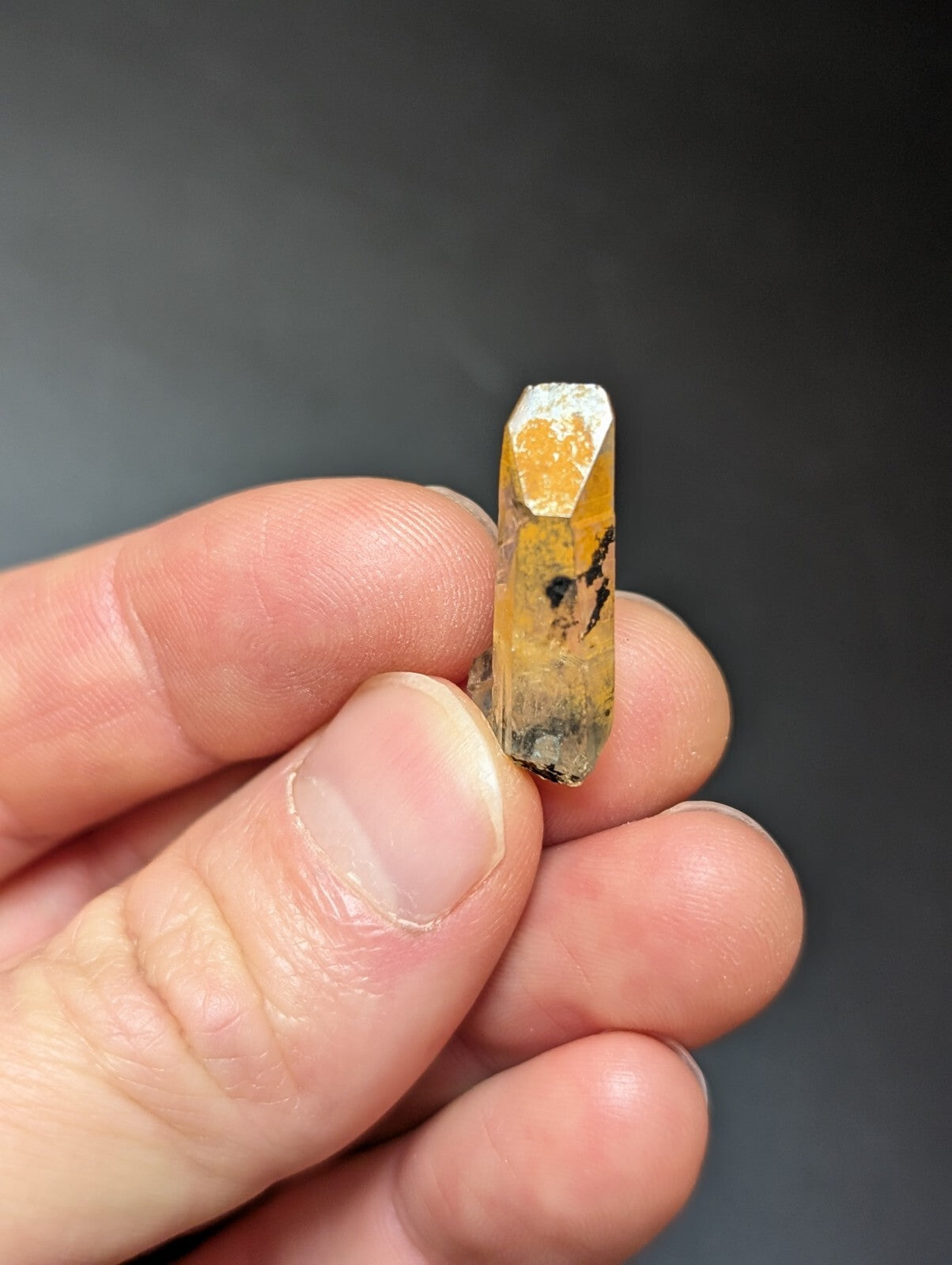 A translucent yellow-orange crystal with black inclusions and a faceted top, held gently between fingers.