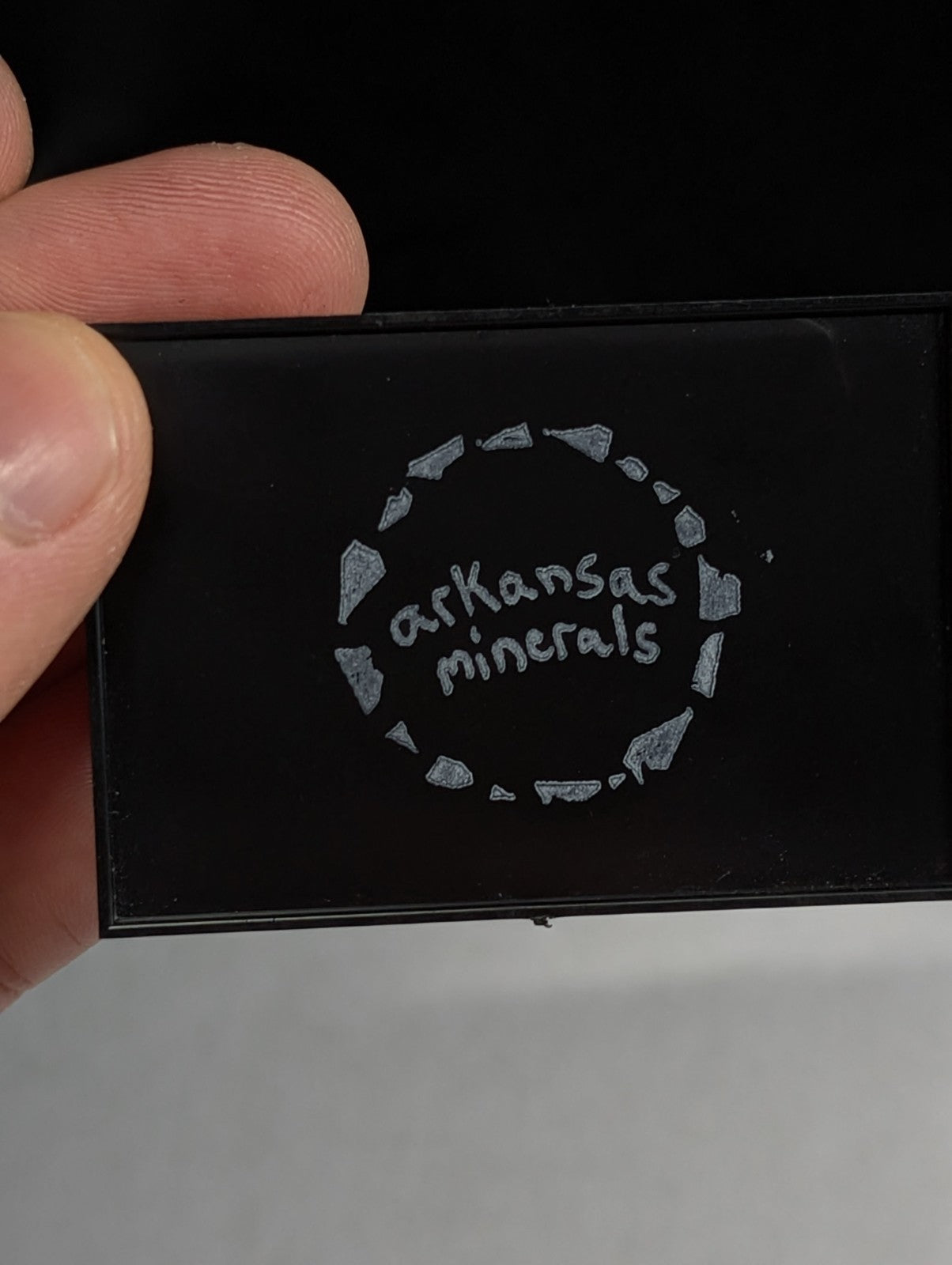 Black rectangular mineral sample with silver engraved logo featuring the text ’arkansas minerals’ surrounded by a circular pattern of stylized rock fragments.
