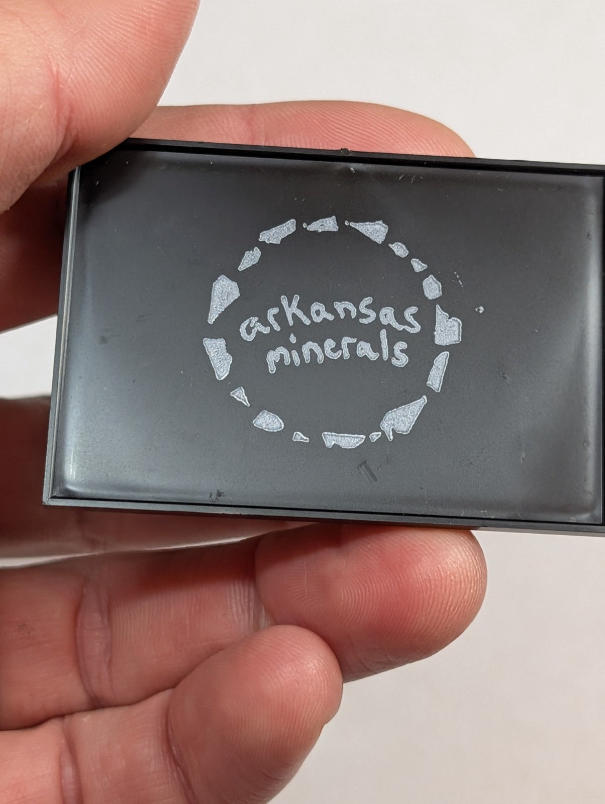 Black rectangular mineral sample box with ’arkansas minerals’ engraved in silver lettering surrounded by a circular pattern of stylized rock fragments.