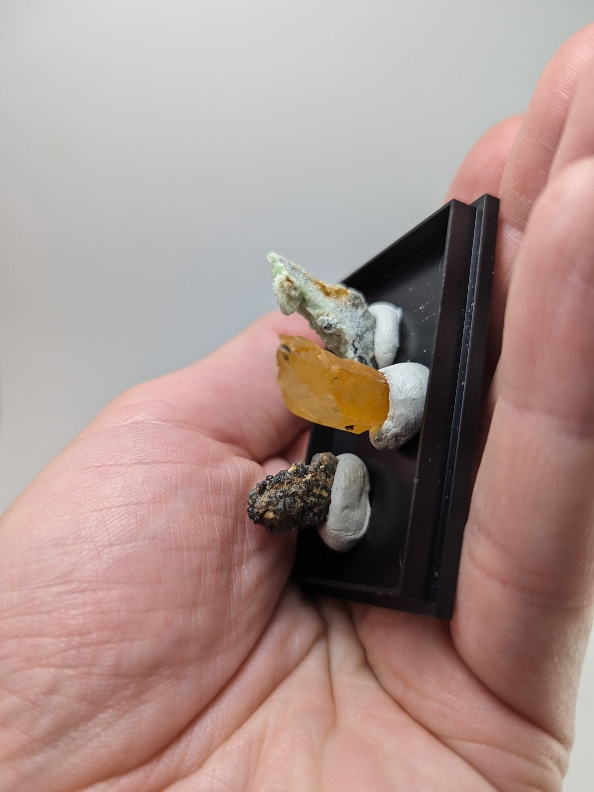 A hand holds a miniature black display case showcasing three distinct mineral specimens, including an amber-colored crystal and grayish-white rock formations.
