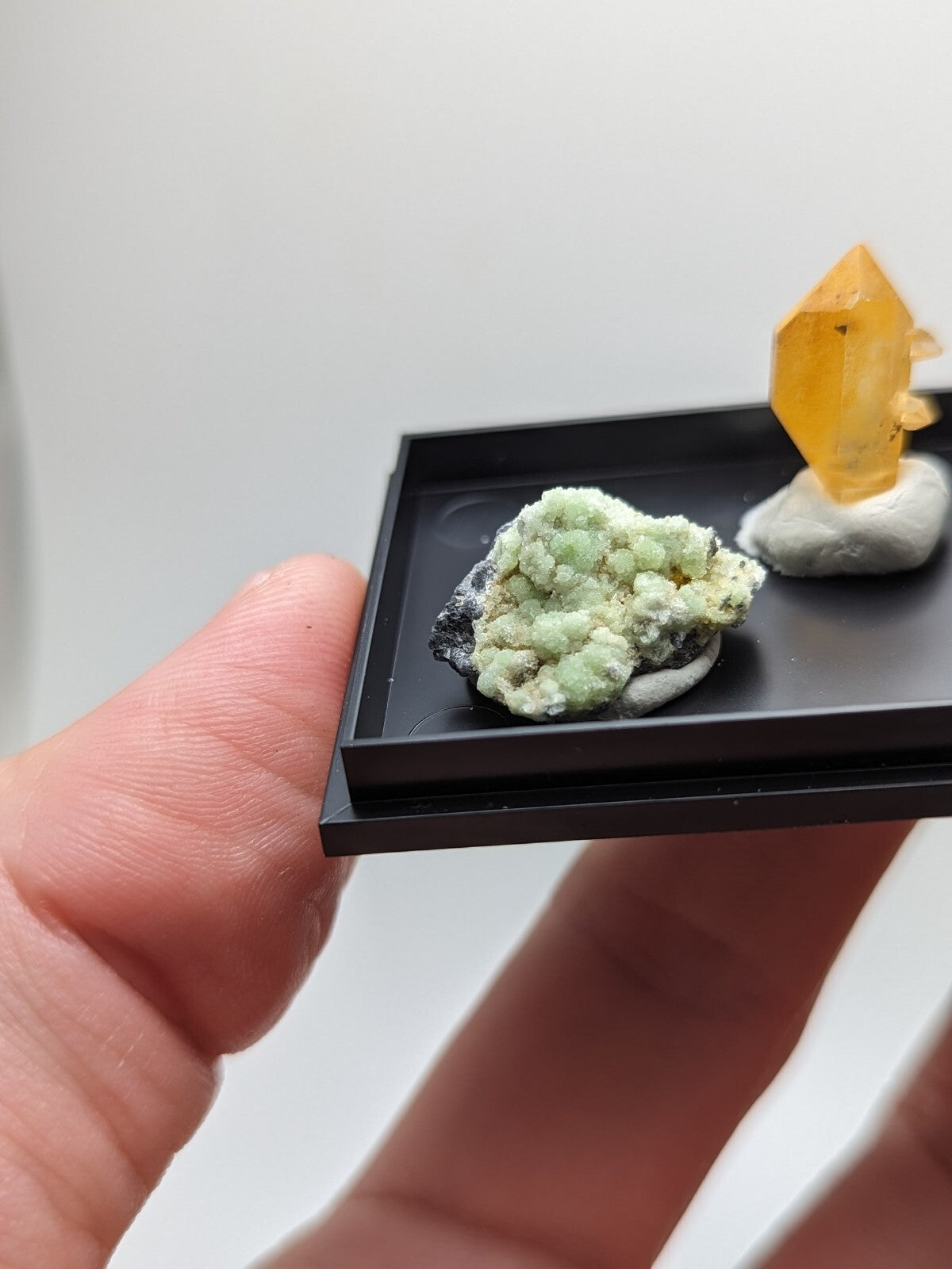 A miniature black display tray holding a green mineral specimen and an amber-colored crystal.
