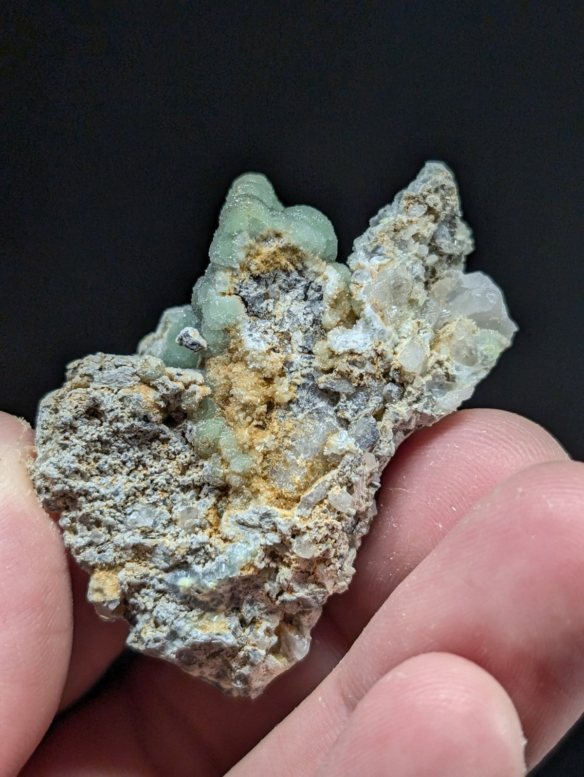 A hand holds a rough, irregularly shaped mineral specimen featuring translucent light green crystals embedded in a matrix of gray and yellowish earthy material.