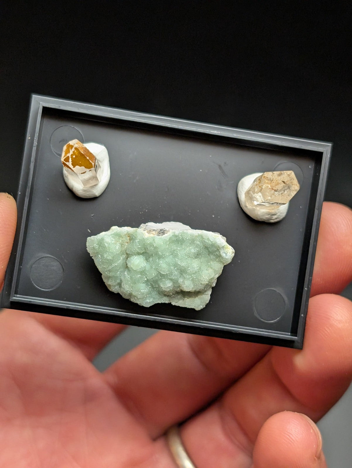 A black rectangular display tray holding three mineral specimens: two small, faceted golden-yellow crystals mounted on white bases and one larger greenish-blue crystalline formation.