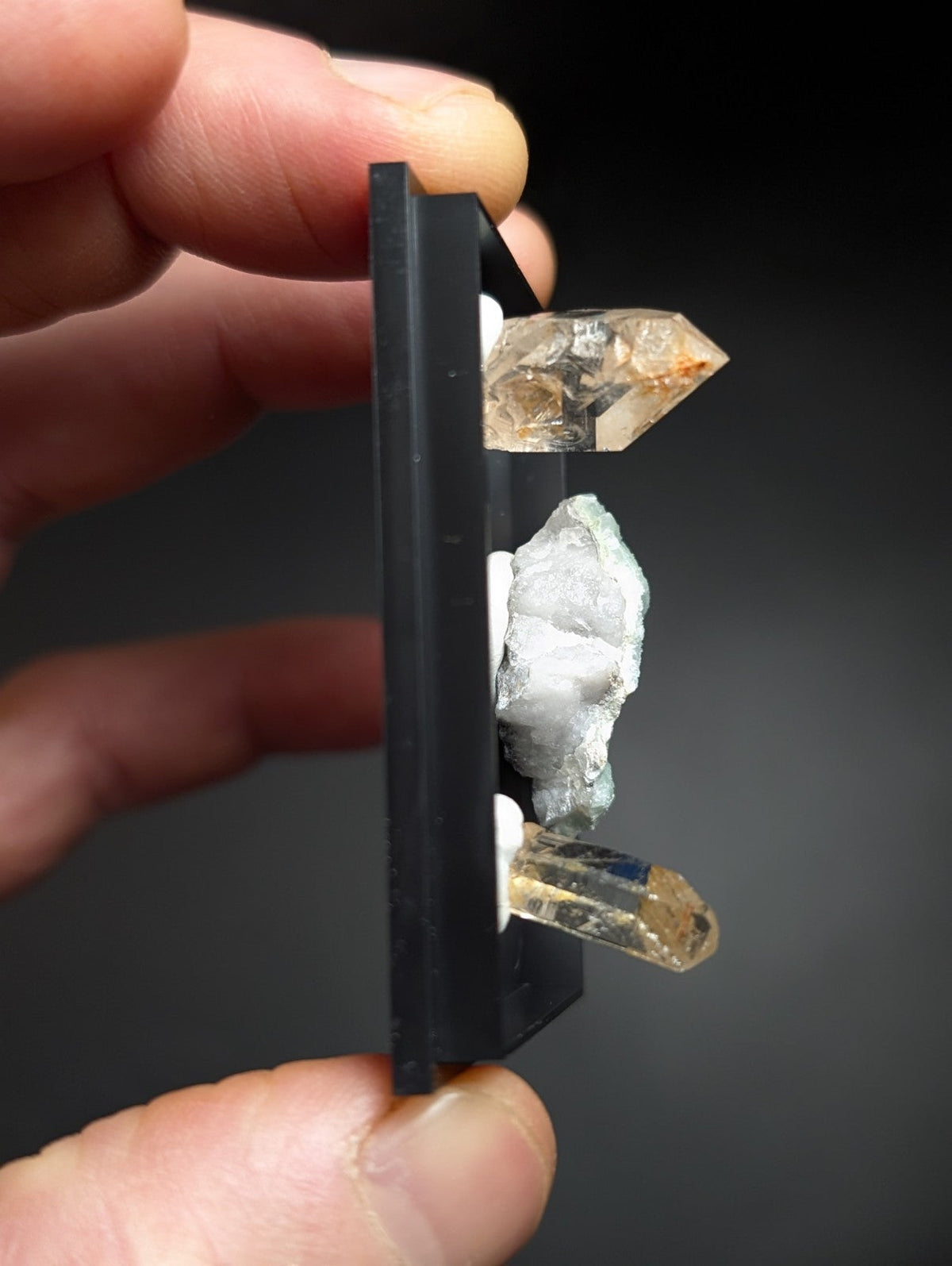 A black display stand holding three distinct quartz crystals, featuring a clear transparent one with amber inclusions at the top and bottom, and a milky white crystalline formation in between.
