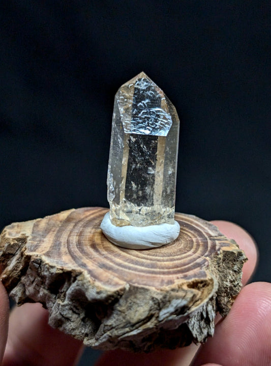 A clear, faceted quartz crystal with a pointed top and natural inclusions sits atop a small white circular base on a rustic wooden slice.