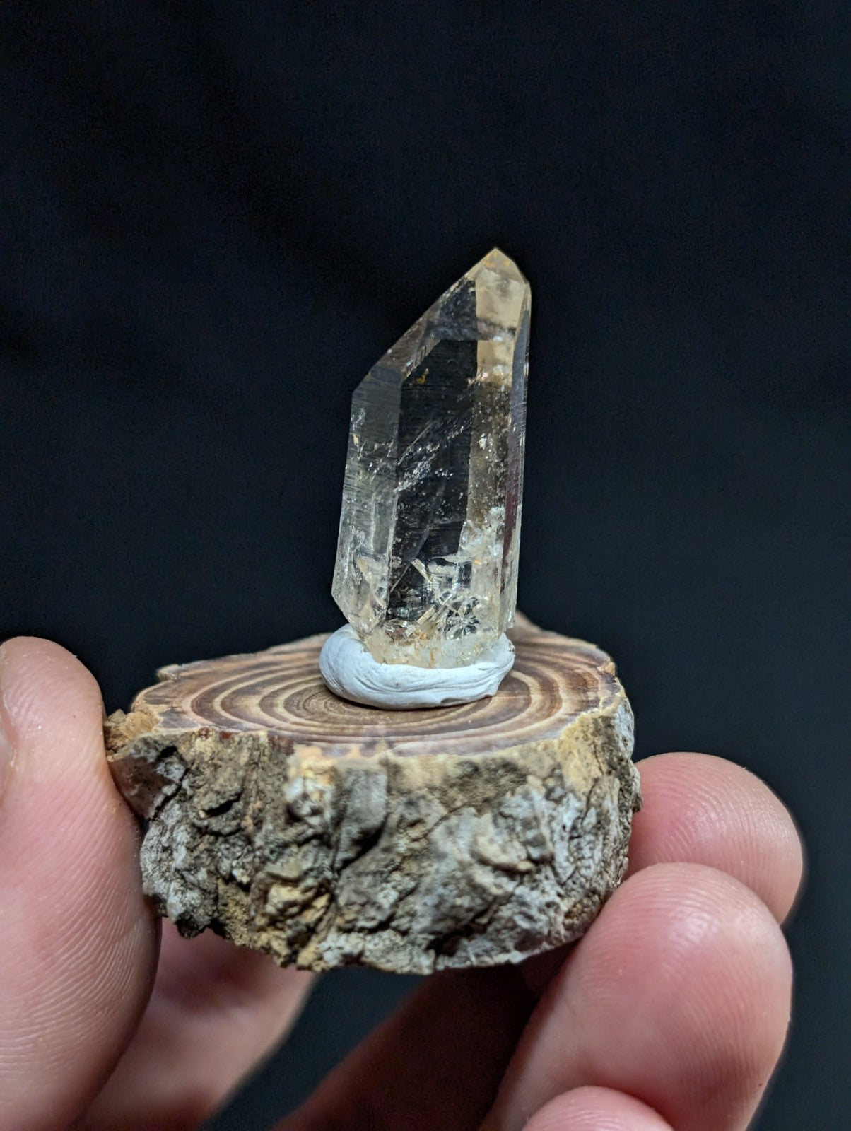 A clear, faceted quartz crystal with subtle golden inclusions stands upright on a rustic wooden slice base.