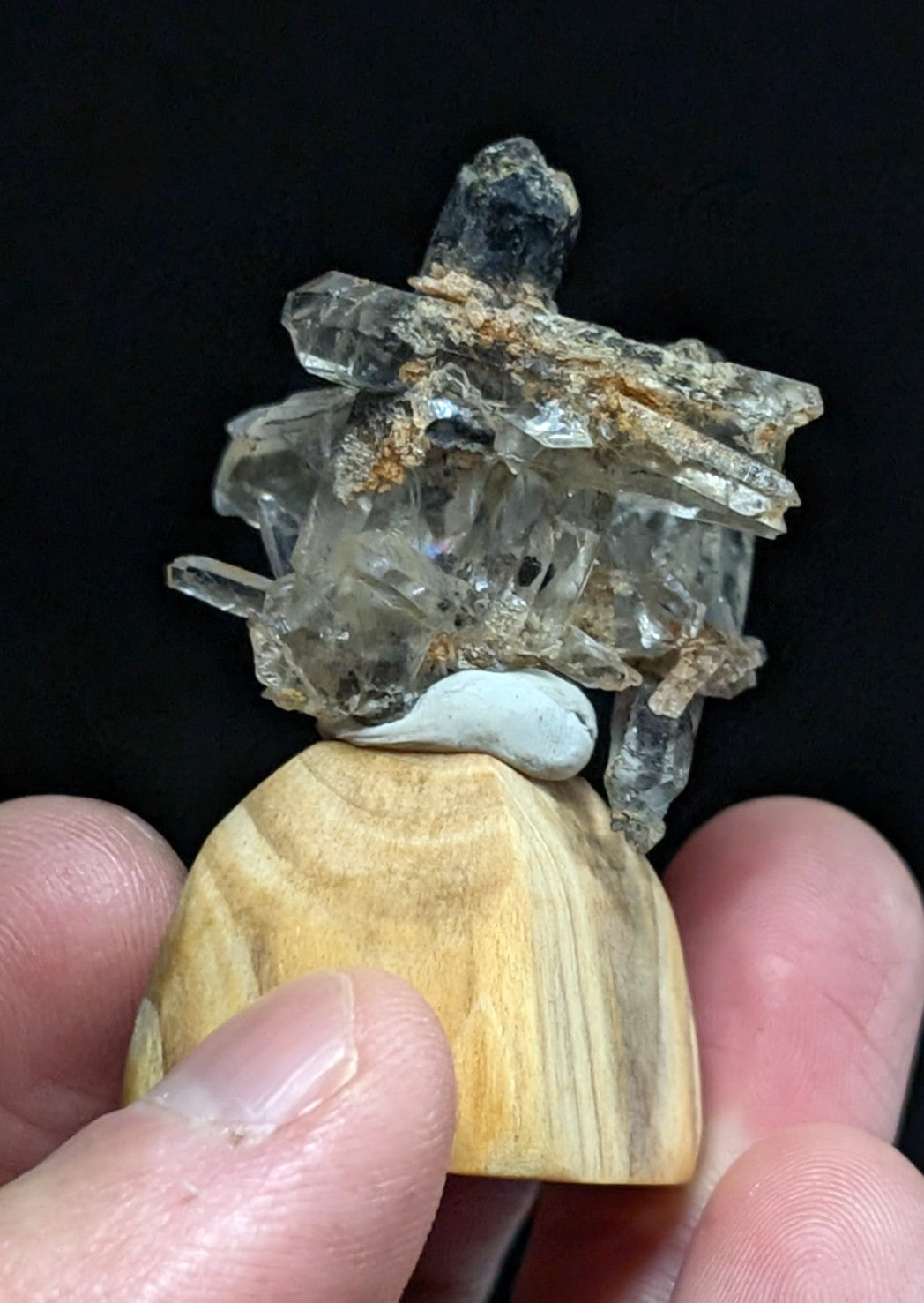 A clear quartz crystal cluster with translucent, faceted formations and earthy brown mineral inclusions rests atop a smooth, light-brown wooden base.