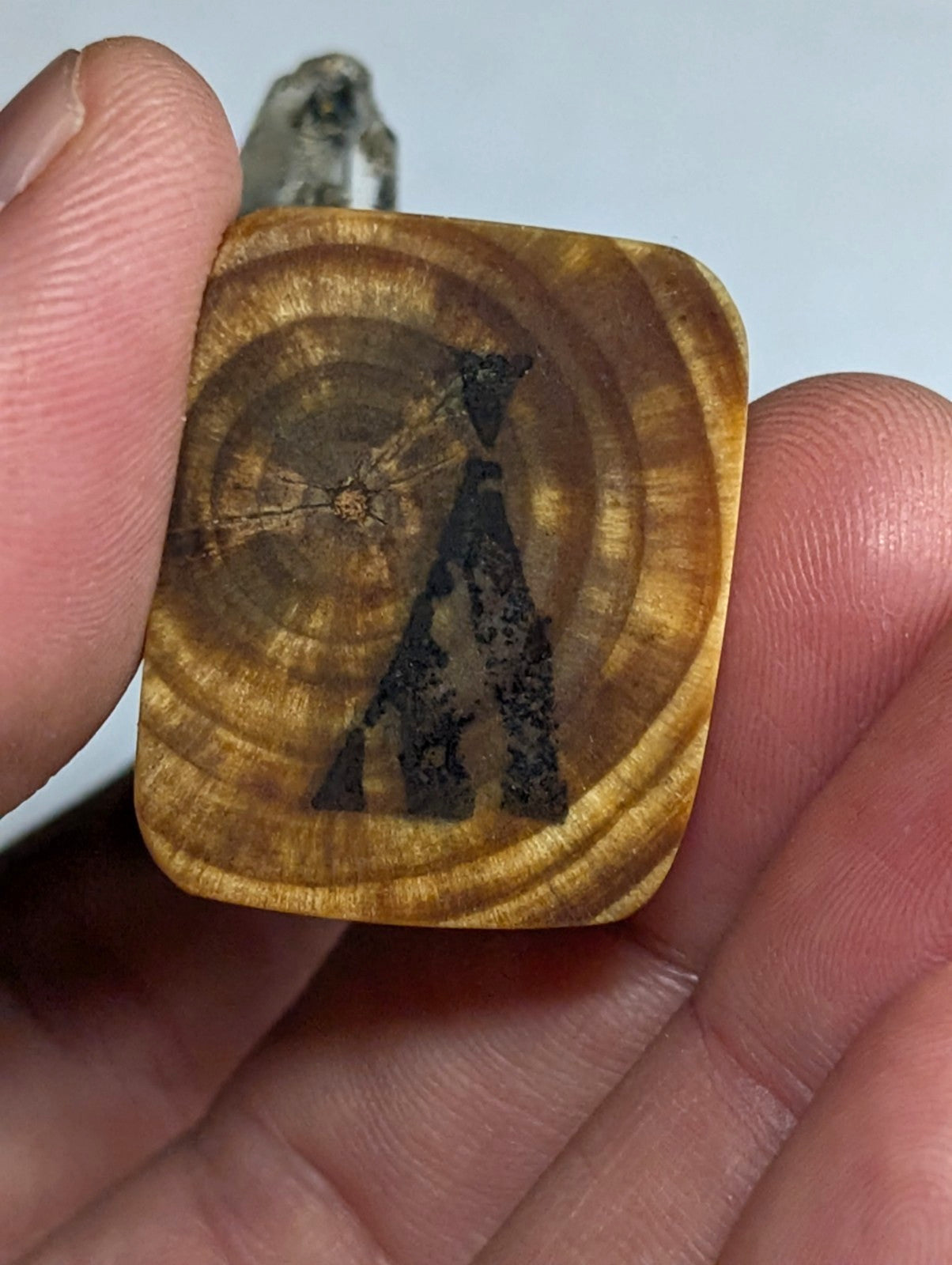 A square-shaped, golden-brown wooden pendant with visible concentric rings and a black silhouette of an abstract figure engraved on its surface.
