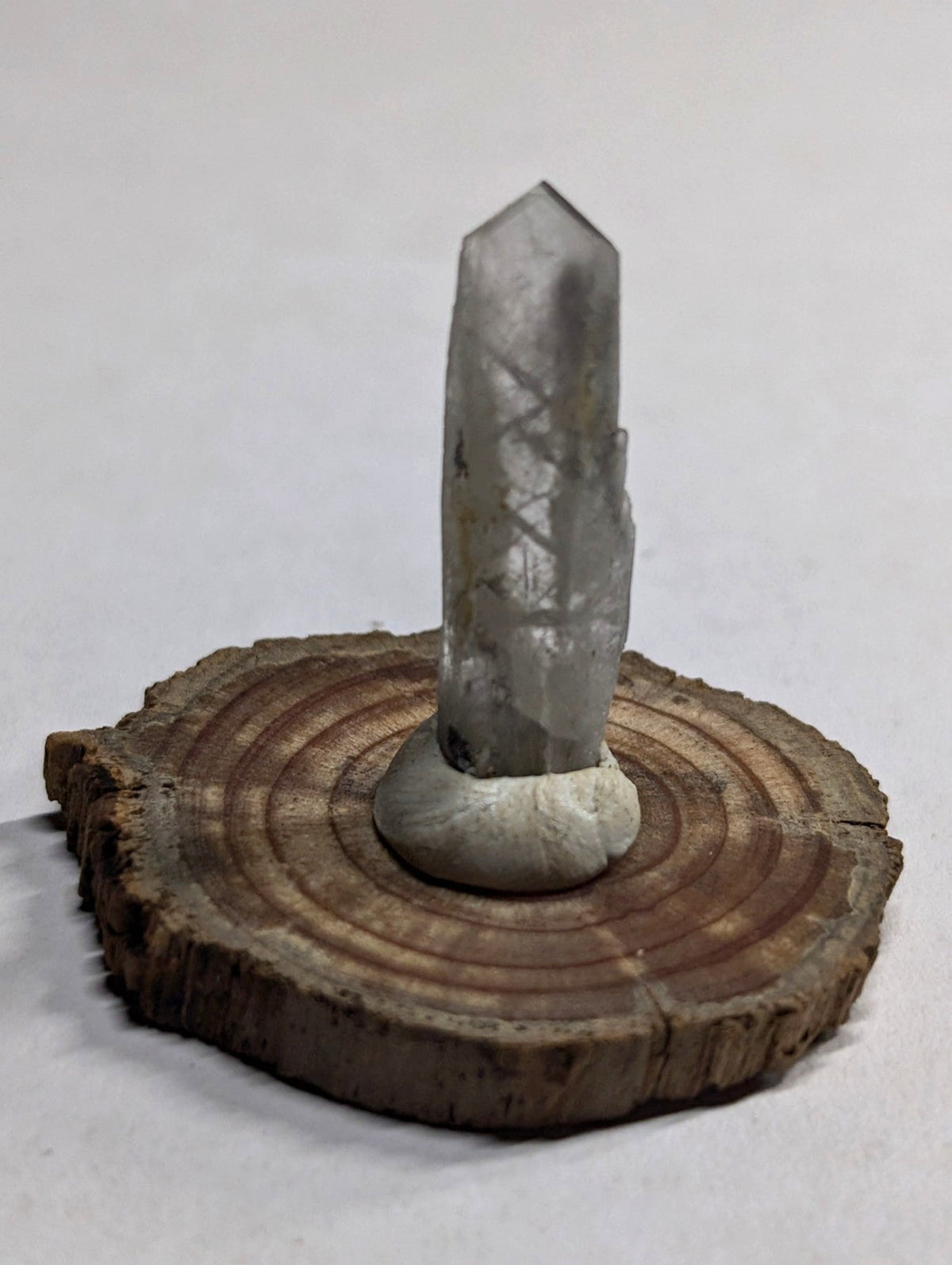 A clear, faceted quartz crystal with subtle internal inclusions stands upright on a circular wooden slice base.