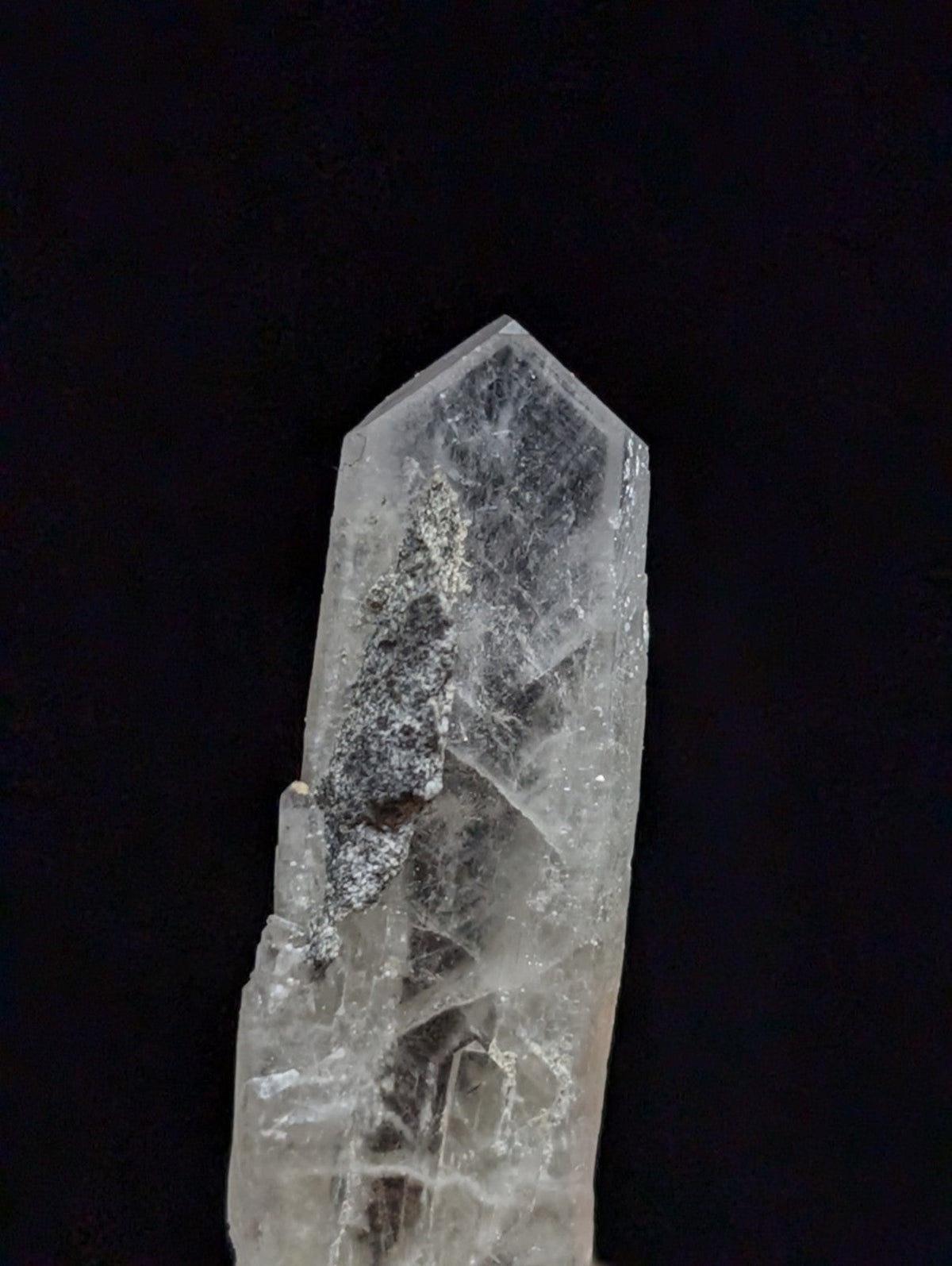 A translucent quartz crystal with a pointed tip and visible internal inclusions, featuring natural striations and subtle gray veining against its clear base.