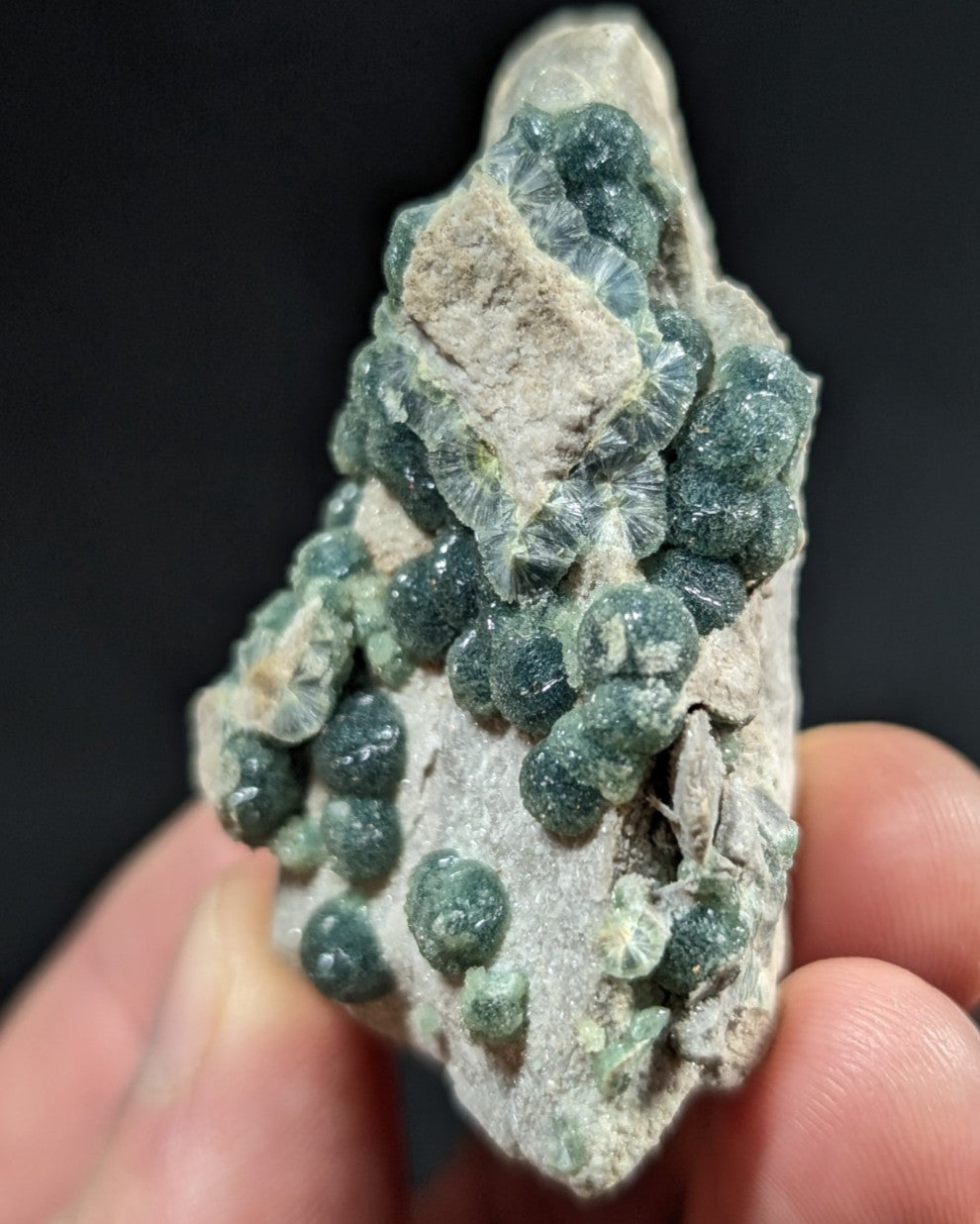 A cluster of dark green, spherical crystals grows densely from a light gray rock matrix.
