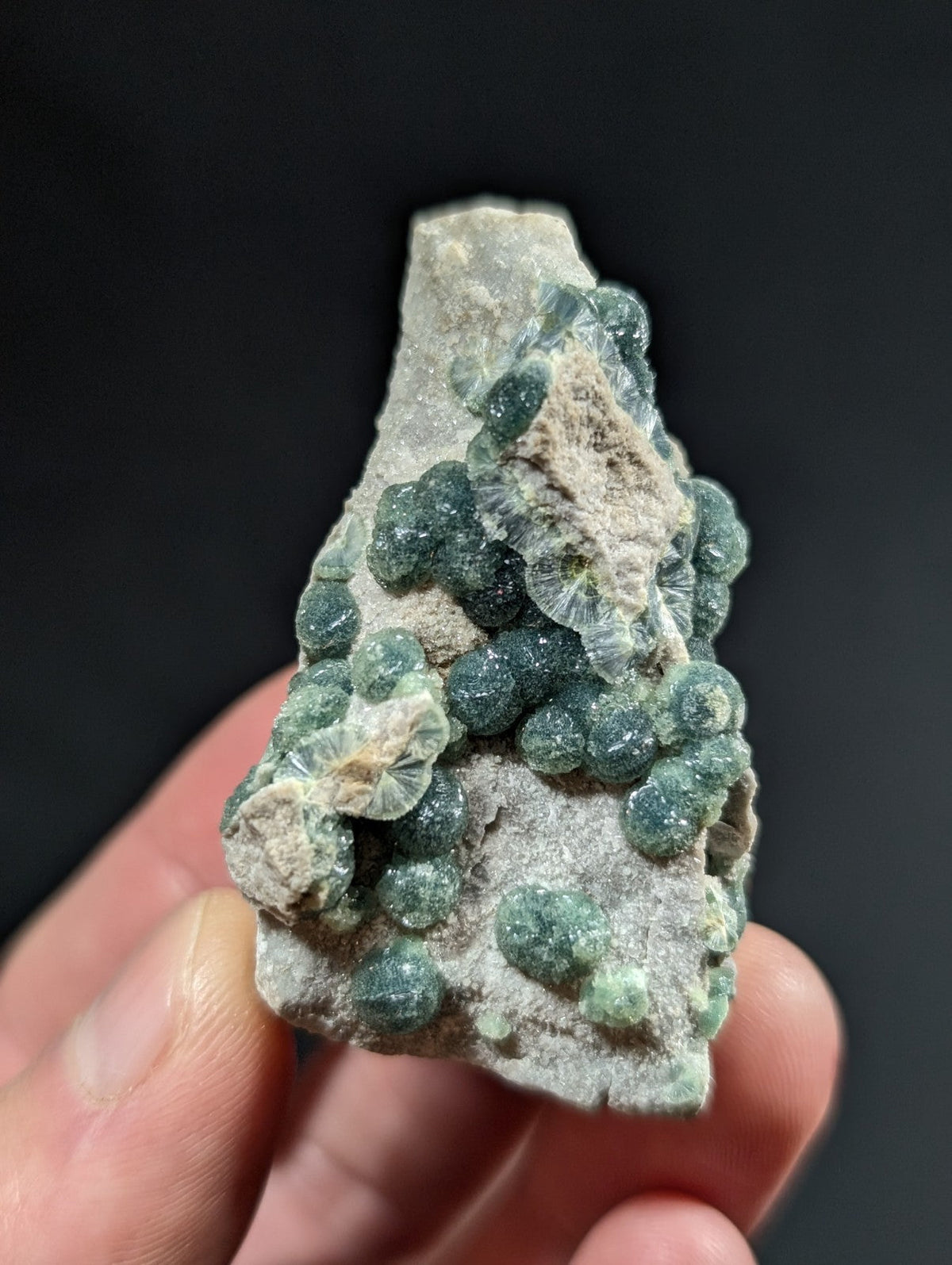 A hand holds a rough, grayish rock specimen featuring clusters of translucent green spherical crystals.
