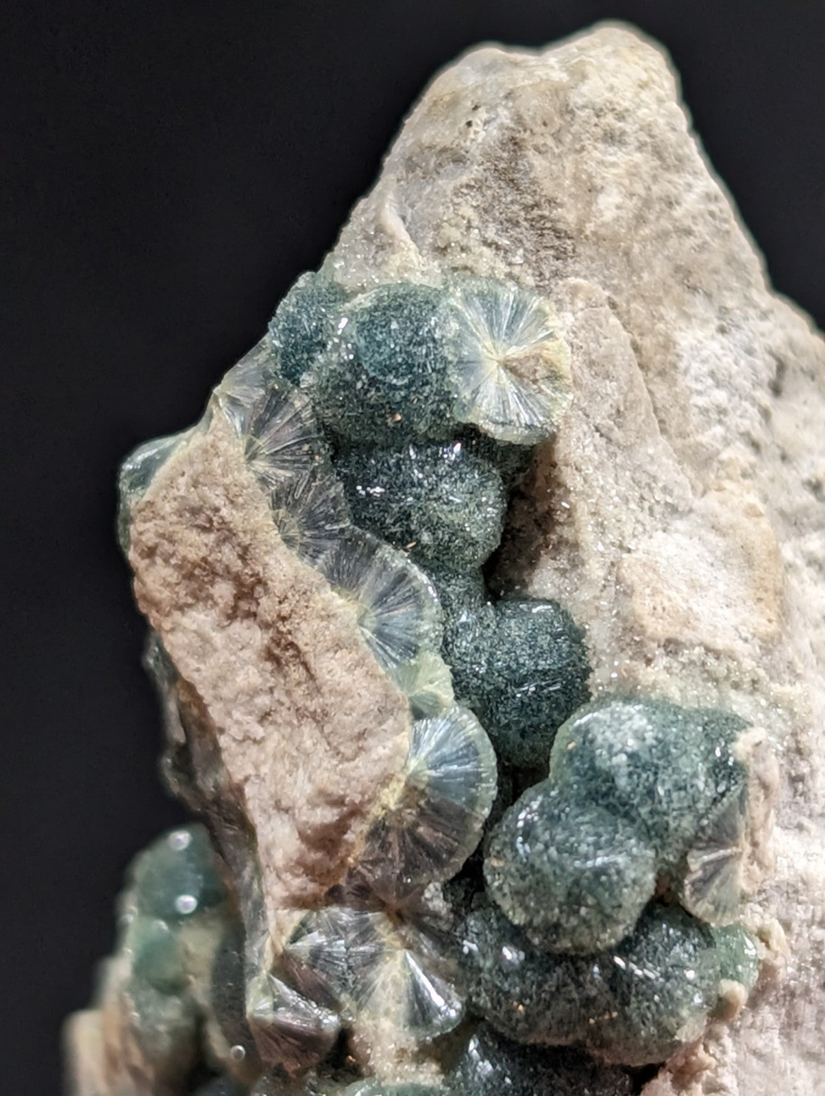 A cluster of translucent, greenish-blue druzy crystals with a radial, fan-like pattern growing from a rough, beige rock matrix.