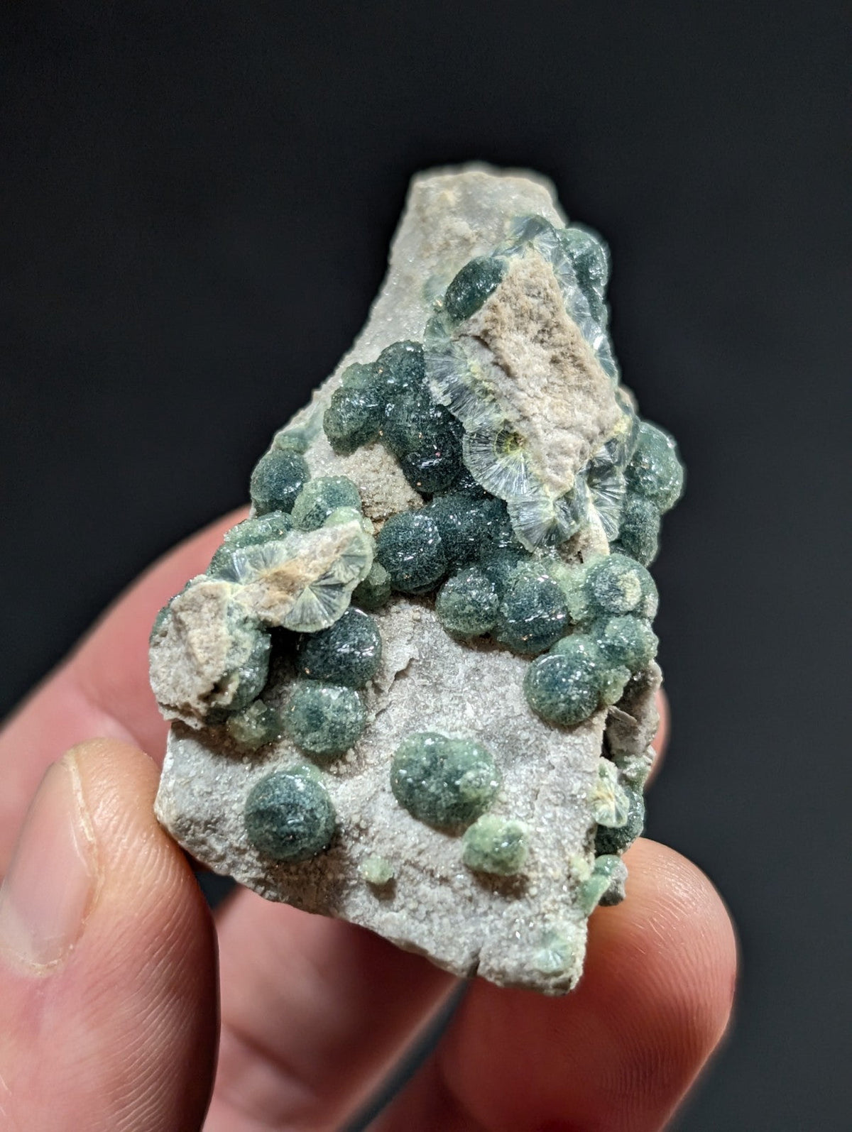 A hand holds a rough, grayish-white rock specimen featuring clusters of small, round, translucent green crystals.