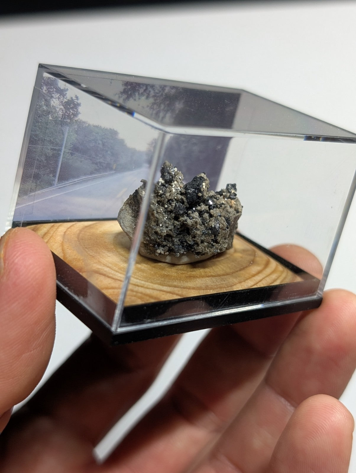 A dark, metallic mineral specimen with a rough, clustered texture is displayed inside a clear acrylic display case resting on a wooden base.