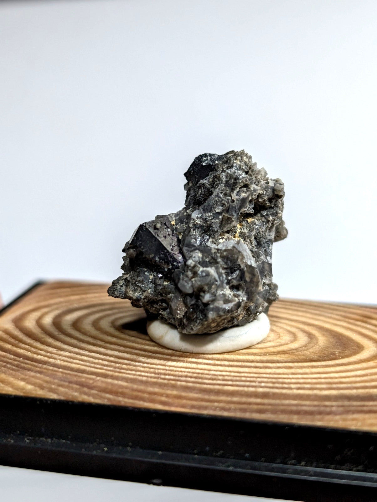 A rugged, dark gray mineral specimen with jagged edges and subtle metallic sheen rests on a circular white base atop a wooden display board.