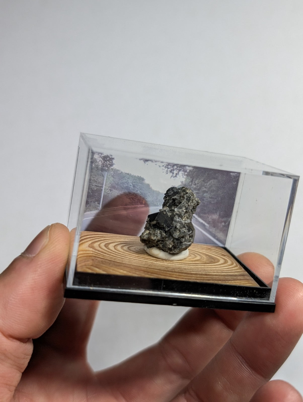 A dark, rough-textured mineral specimen displayed in a clear acrylic display case with a wooden base.