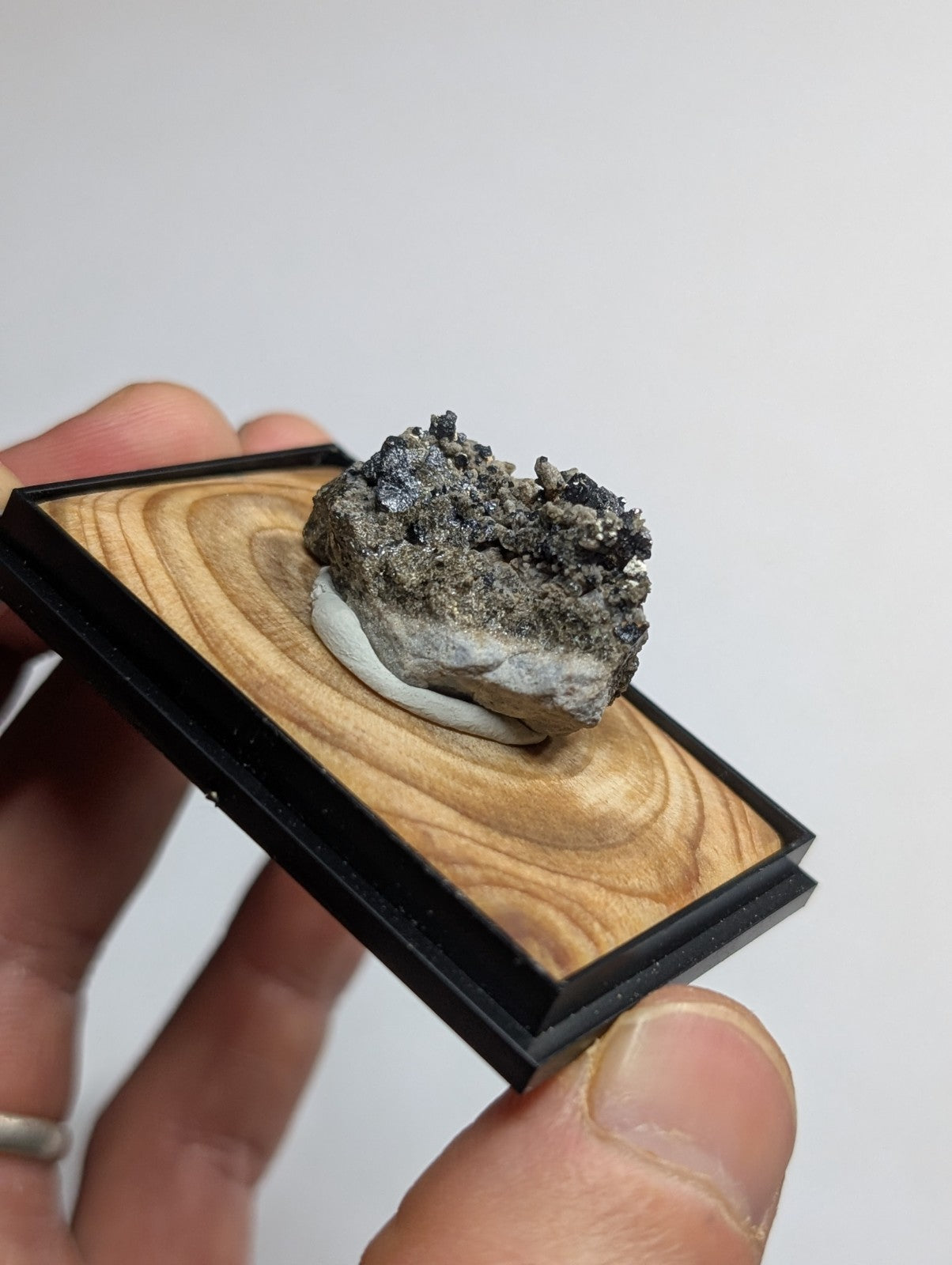 A dark gray, textured mineral specimen with a rough, crystalline surface rests on a light wood display base framed in black.