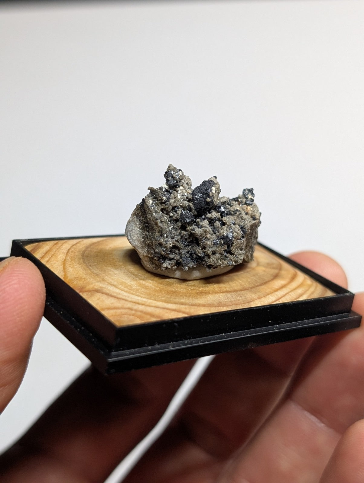 A gray and black mineral specimen with a rough, crystalline texture displayed on a wooden base within a sleek black frame.