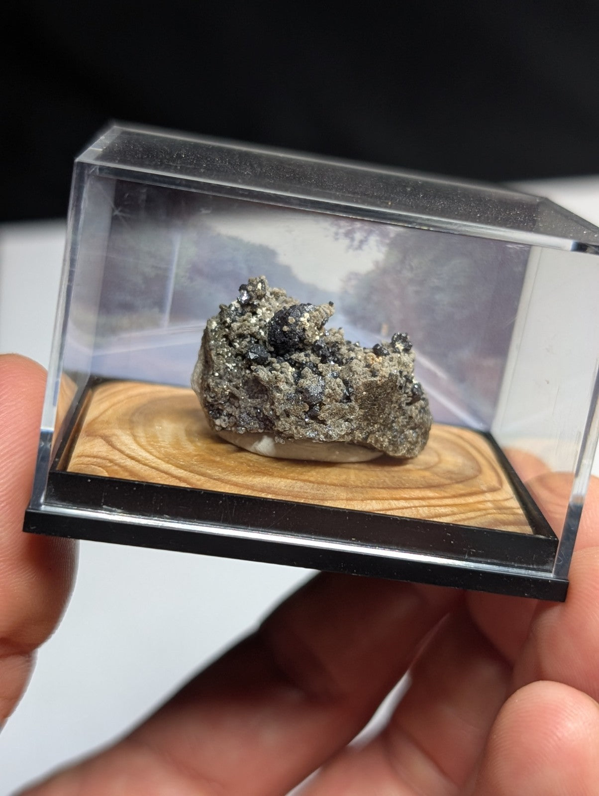 A metallic, textured mineral specimen with a mix of silver and dark gray hues is displayed inside a clear acrylic display case.