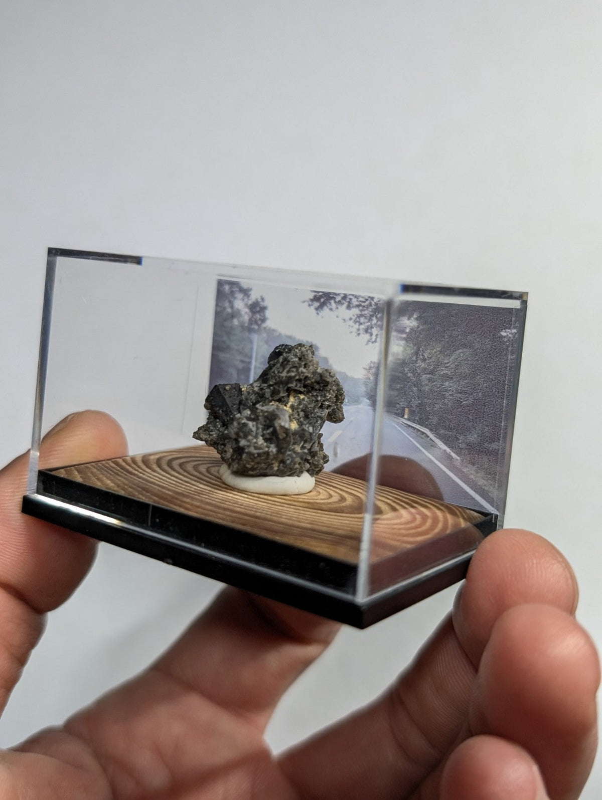 A dark, rough-textured mineral specimen displayed in a clear acrylic display case with a wooden base.