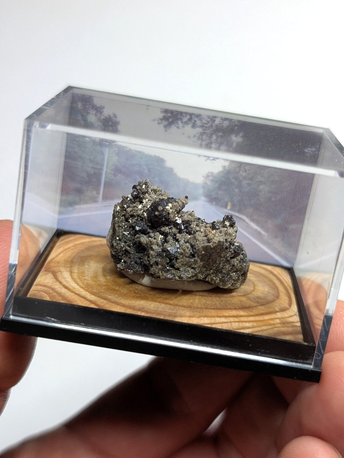 A textured, metallic gray mineral specimen with dark inclusions sits displayed inside a clear acrylic showcase.