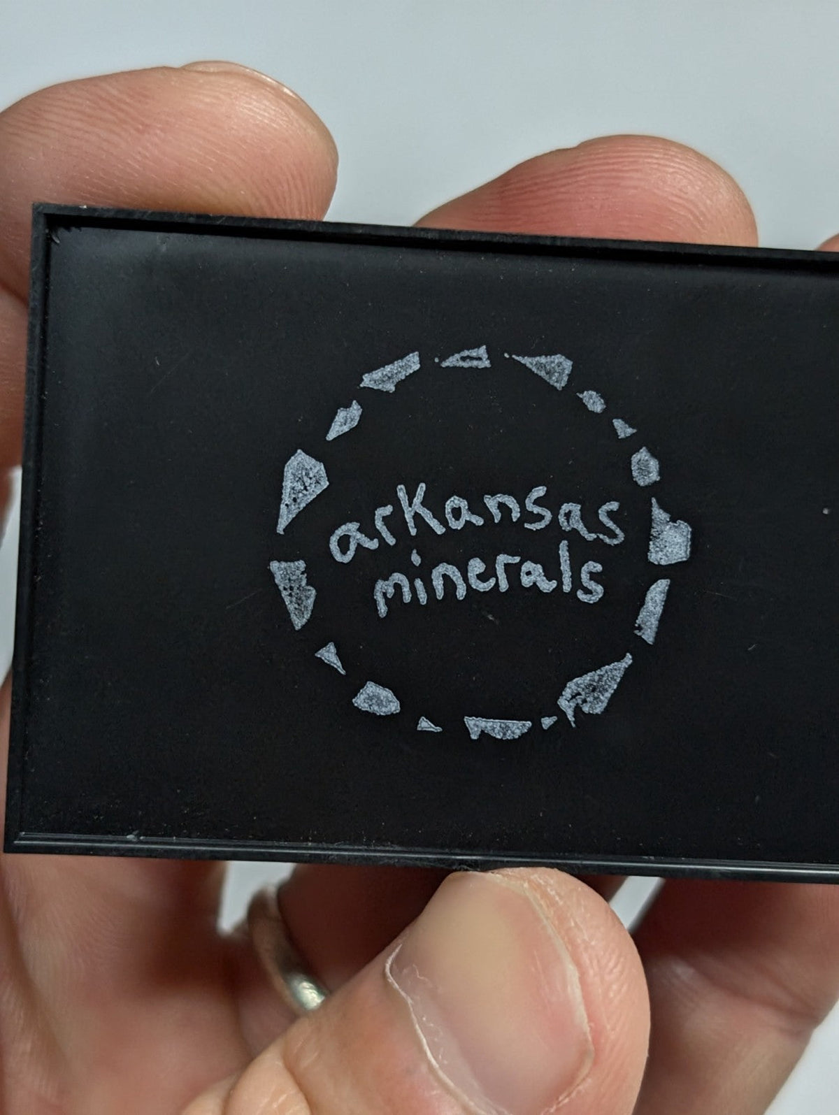 Black rectangular compact with silver ’arkansas minerals’ logo and diamond-shaped border design.