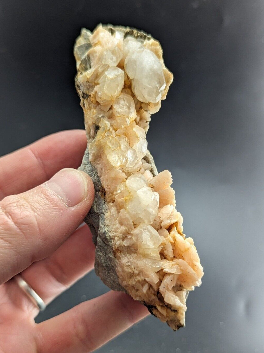 A hand holds a quartz crystal cluster with translucent, pale yellow and white crystals growing from a rough, grayish rock base.