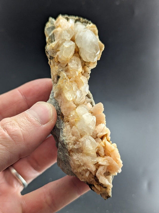 A hand holds a quartz crystal cluster with translucent, pale yellow and white crystals growing from a rough, grayish rock base.