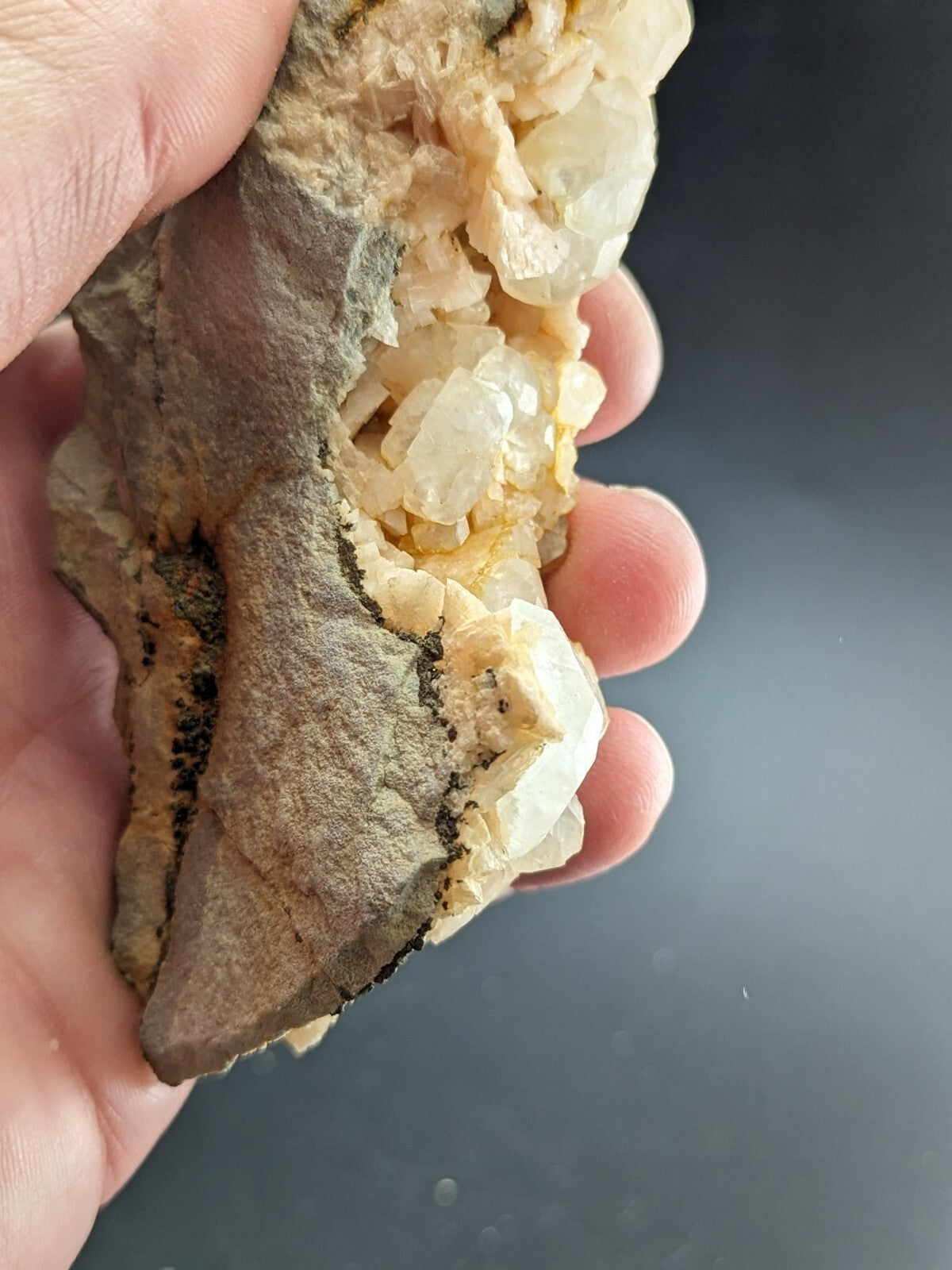 A hand holds a geological specimen featuring translucent, crystalline formations with pale yellow and white hues emerging from a rough, earthy gray matrix.