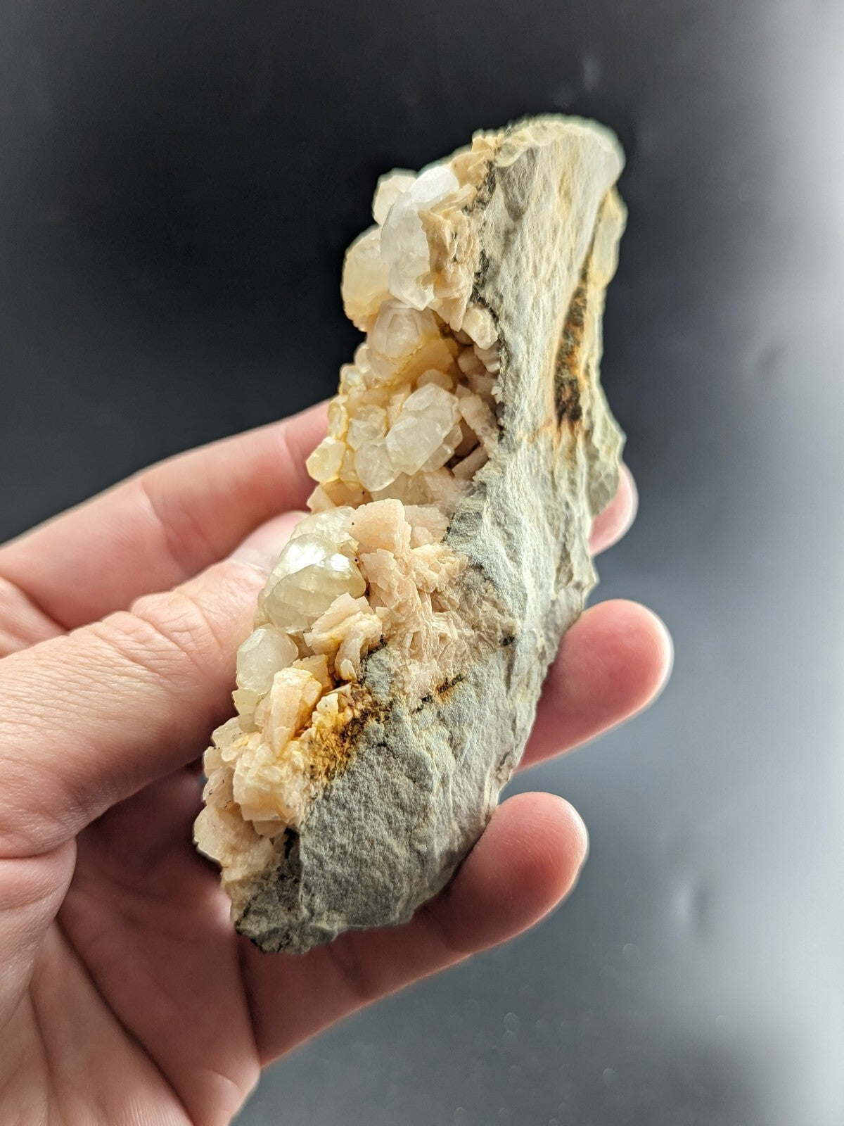 A hand holds a geode with translucent, pale yellowish crystals growing from its rough gray rock matrix.