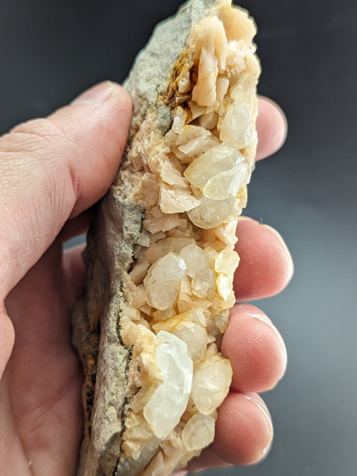 A hand holds a geological specimen featuring translucent, pale yellow to white quartz crystals growing from a rough, grayish rock matrix.
