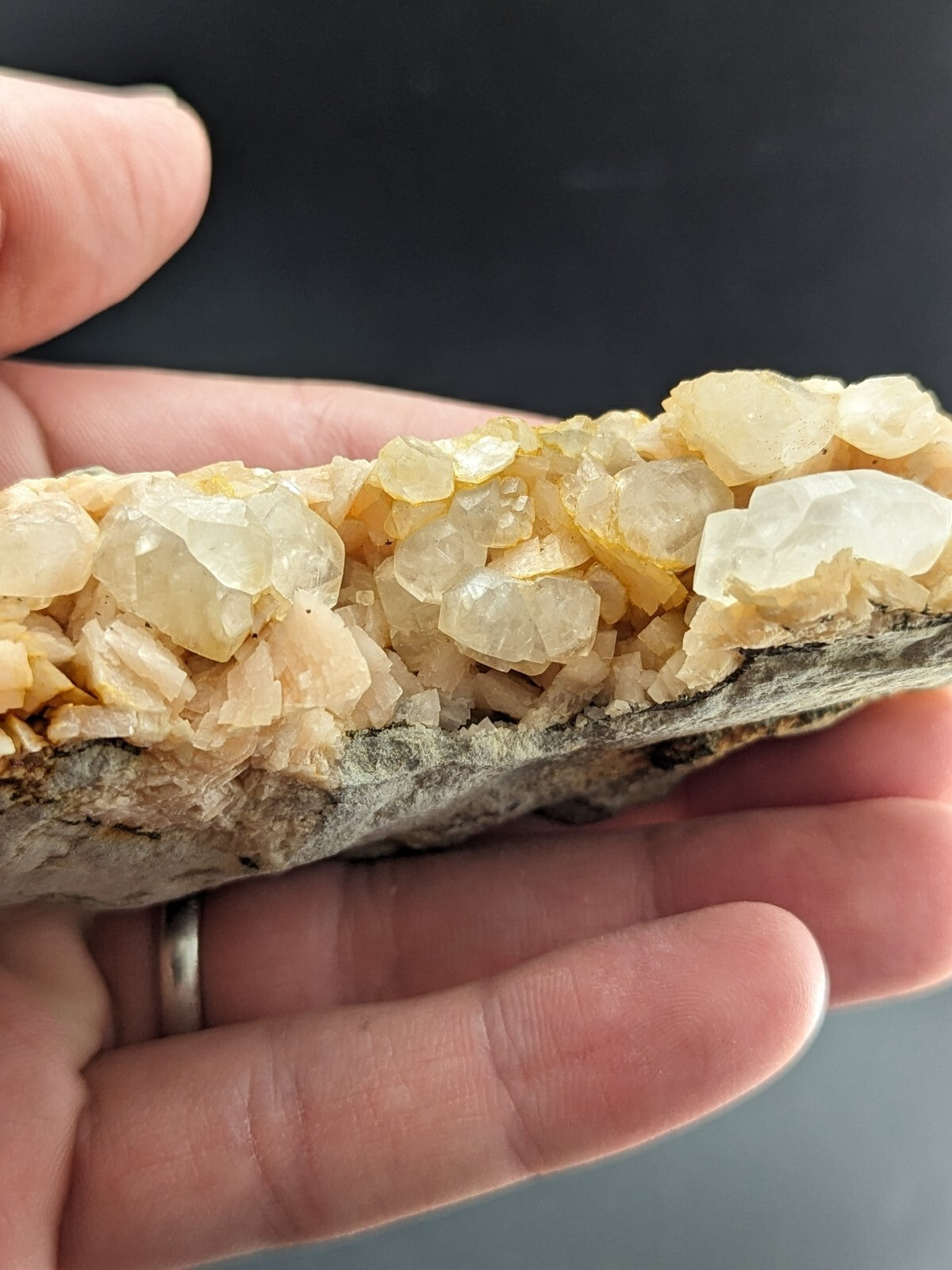 A hand holds a geological specimen featuring clusters of translucent, pale yellow to white crystals growing from a dark gray rock base.
