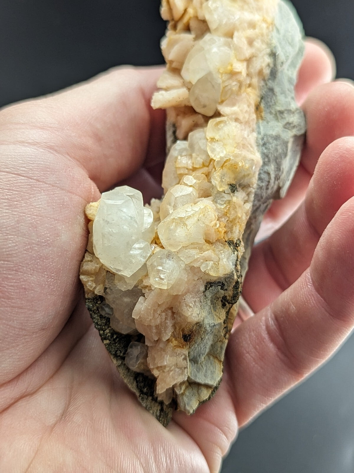 A hand holds a geological specimen featuring translucent, pale yellow and white crystalline formations growing from a dark gray rock base.