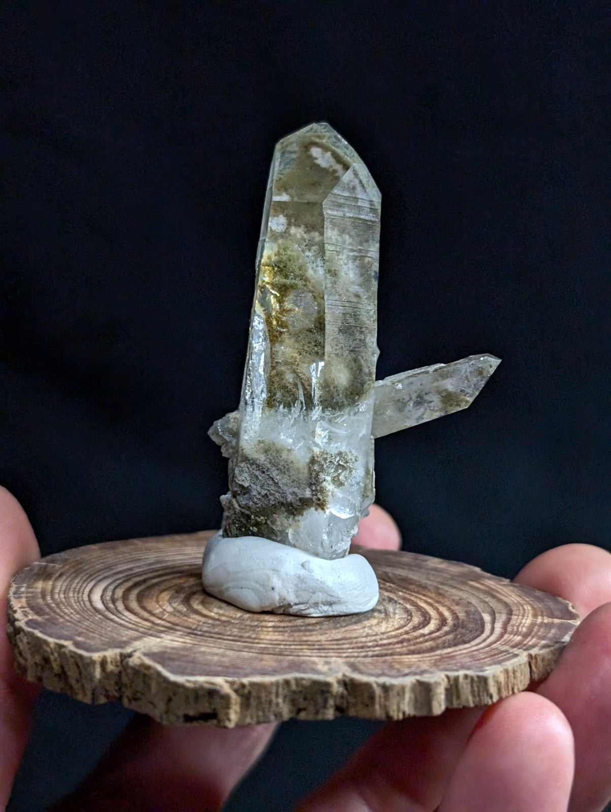 A translucent, pale green quartz crystal cluster with golden inclusions and sharp facets stands upright on a smooth white stone base atop a rustic wooden slice.