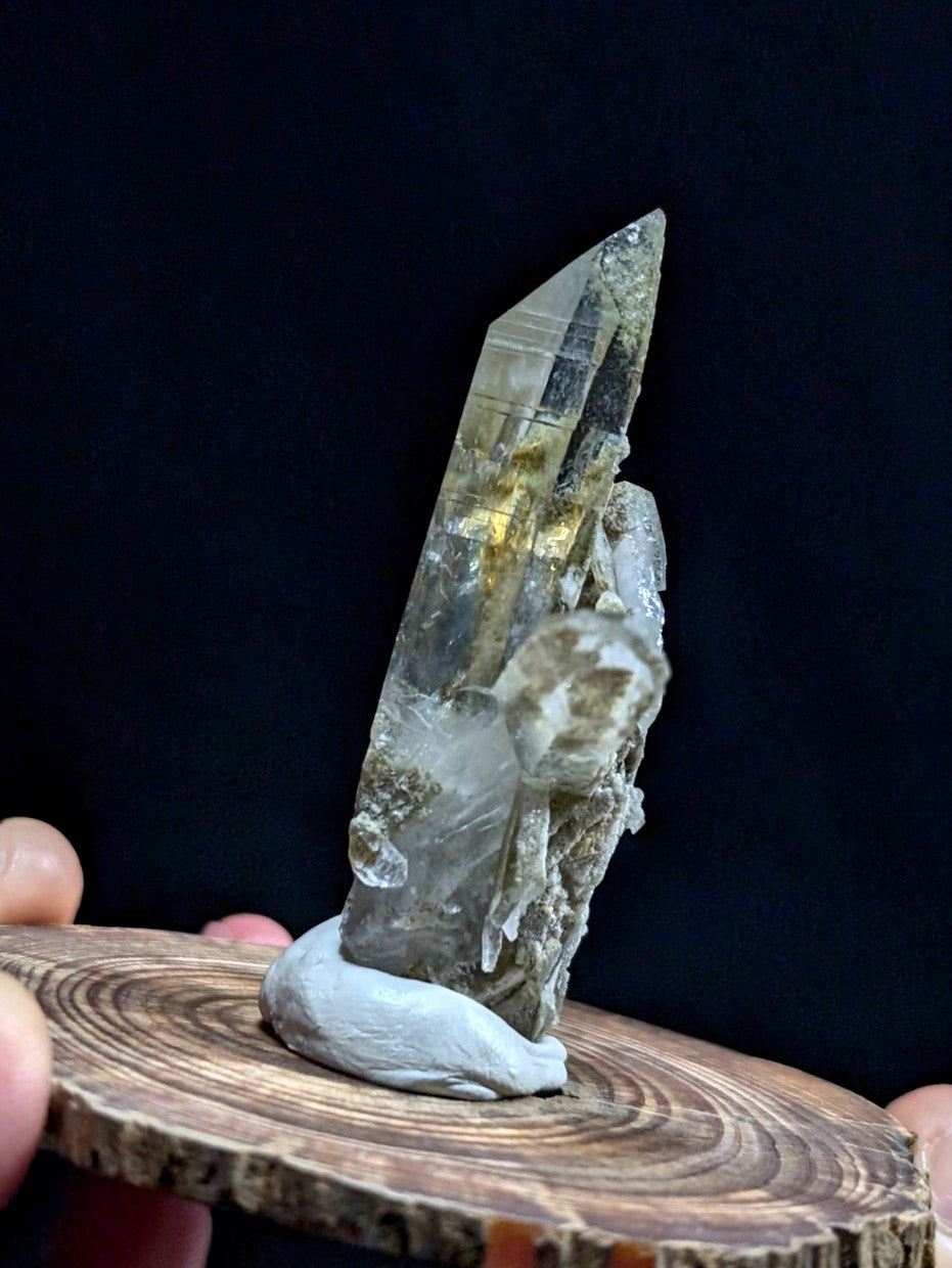 A translucent, pointed quartz crystal with golden inclusions and a rough, earthy base mounted on a wooden slice.