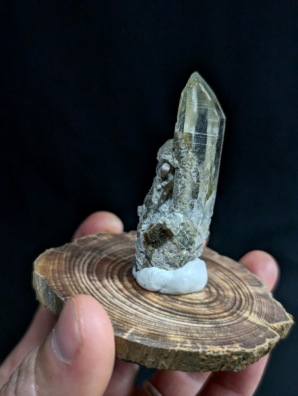 A translucent green quartz crystal with a pointed tip stands upright on a rustic wooden slice, its base secured by a small white resin cap.